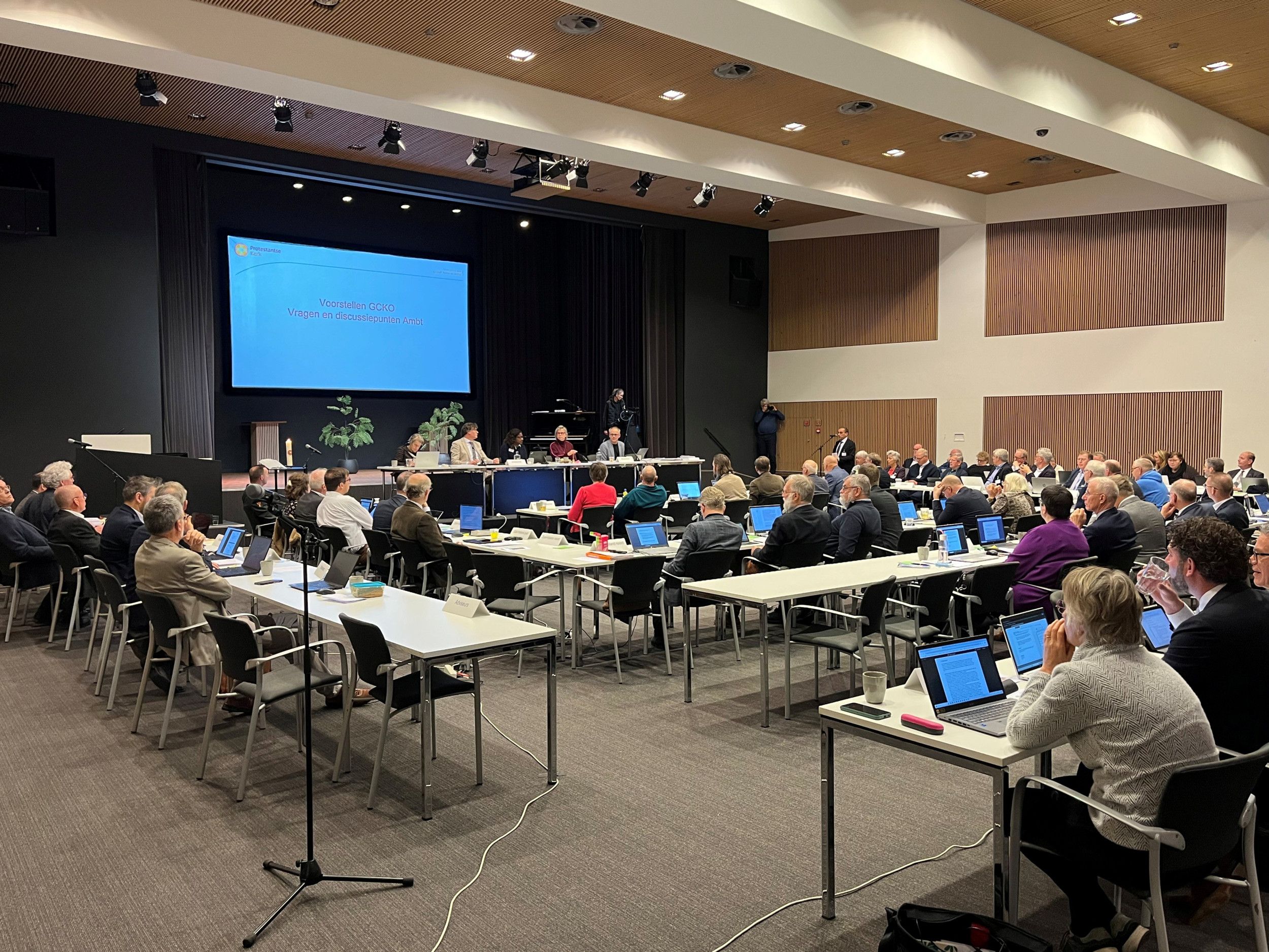 Vergaderzaal met tafels waarachter mannen en vrouwen zitten.