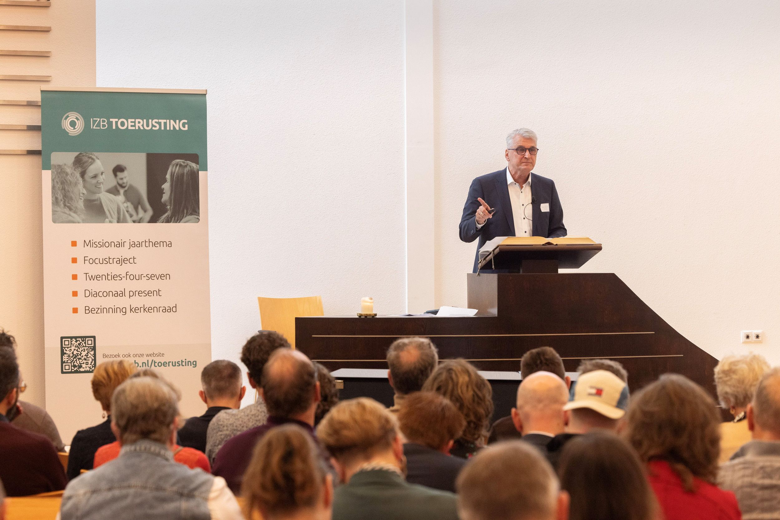 Man met colbert achter een katheder. Links een informatiepaneel van IZB toerusting.