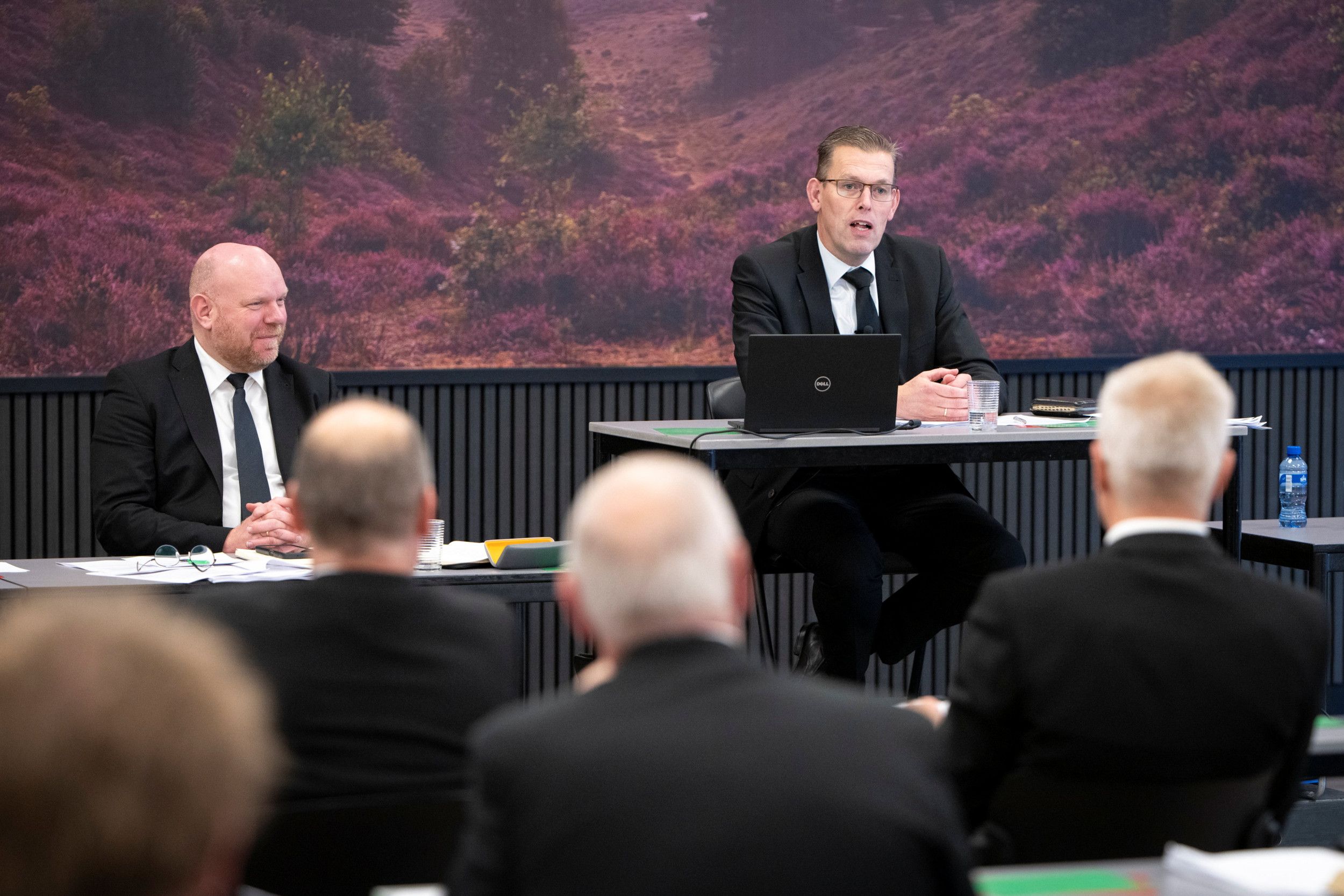 Dominee Diepenbroek en dominee Mulder, twee mannen in zwart pak, zitten achter tafels; voor hen andere mannen.