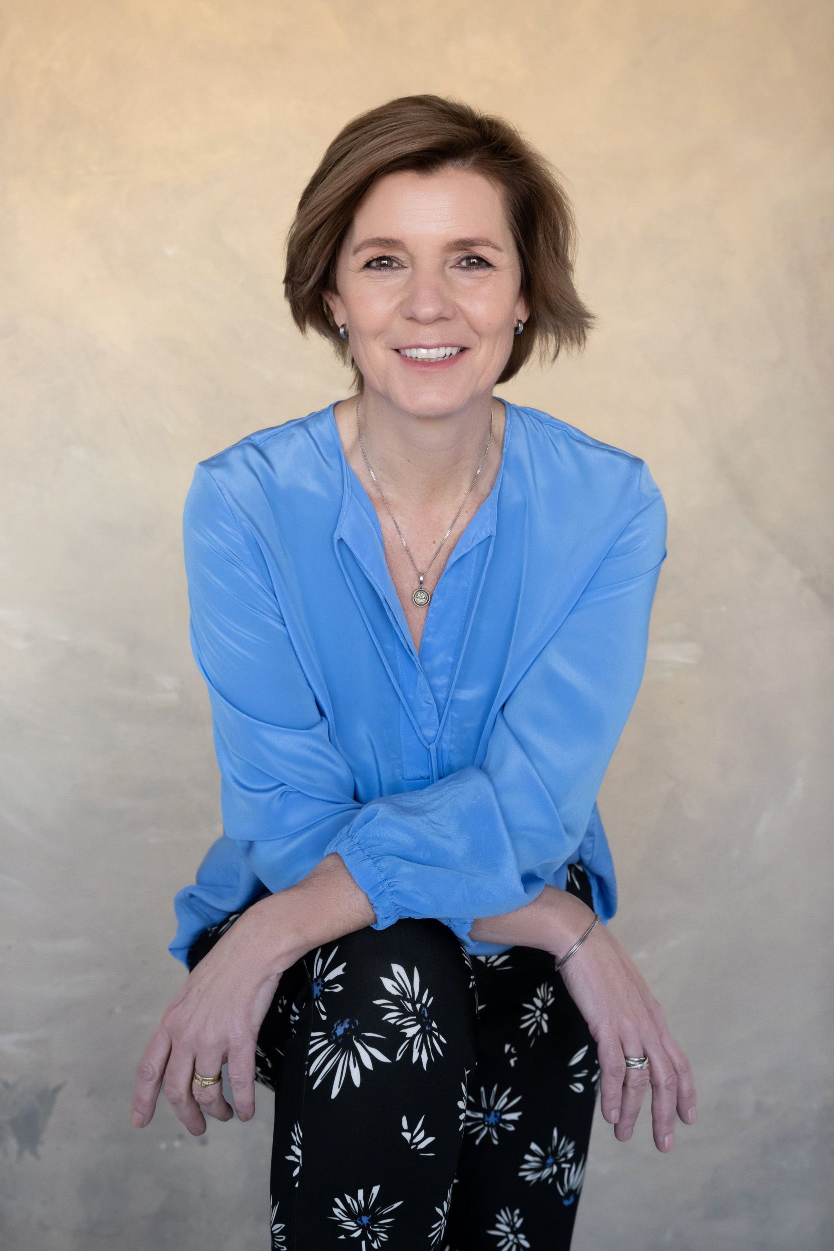 Vrouw met kort bruin haar, zittend, blauwe blouse en zwart gebloemde broek