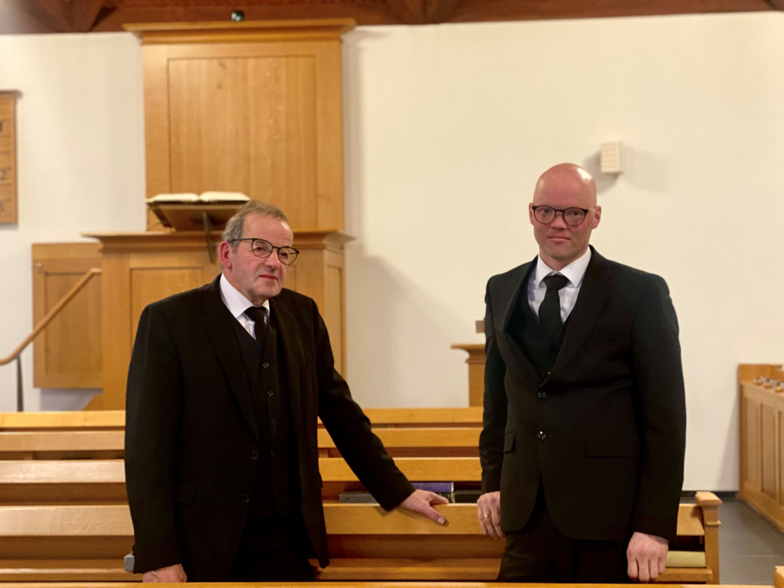 Twee mannen in driedelig zwart pak poseren in een kerkzaal voor een portretfoto.