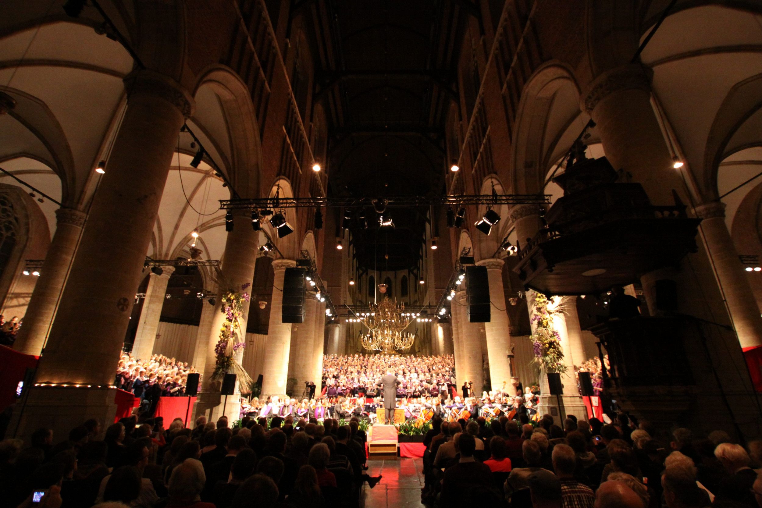 In de Pieterskerk in Leiden staan voor het orgel jongeren op een enorm, oplopend podium opgesteld. Daarvoor het orkest met dirigent. Erboven gouden kroonluchters en links en rechts pilaren. De kerk is goed gevuld met luisteraars.