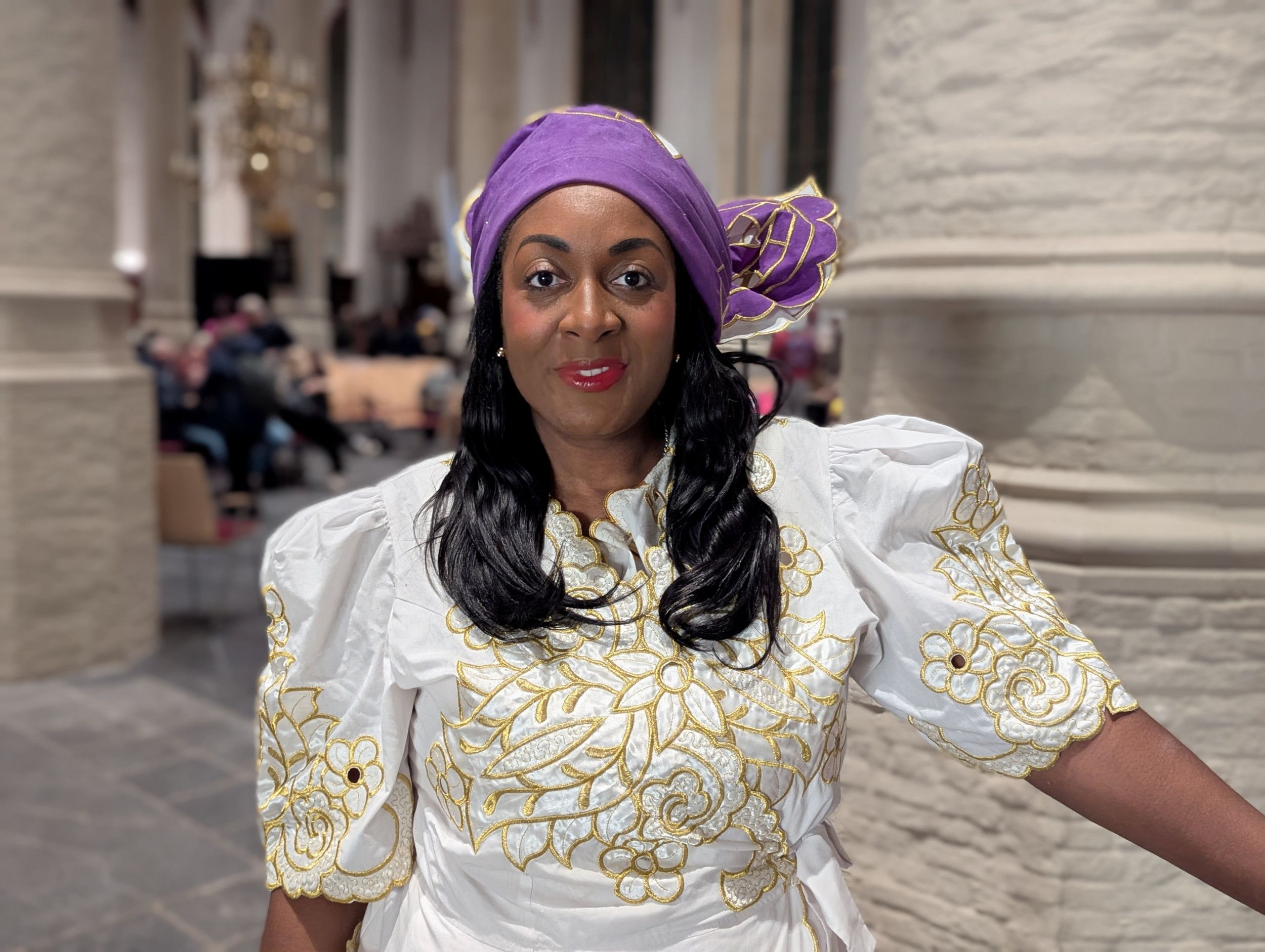 Een donkere vrouw met lang, zwart haar in traditionele Surinaamse kleding: een witte jurk met geborduurde, gouden bloemen en een paarse hoed/muts.