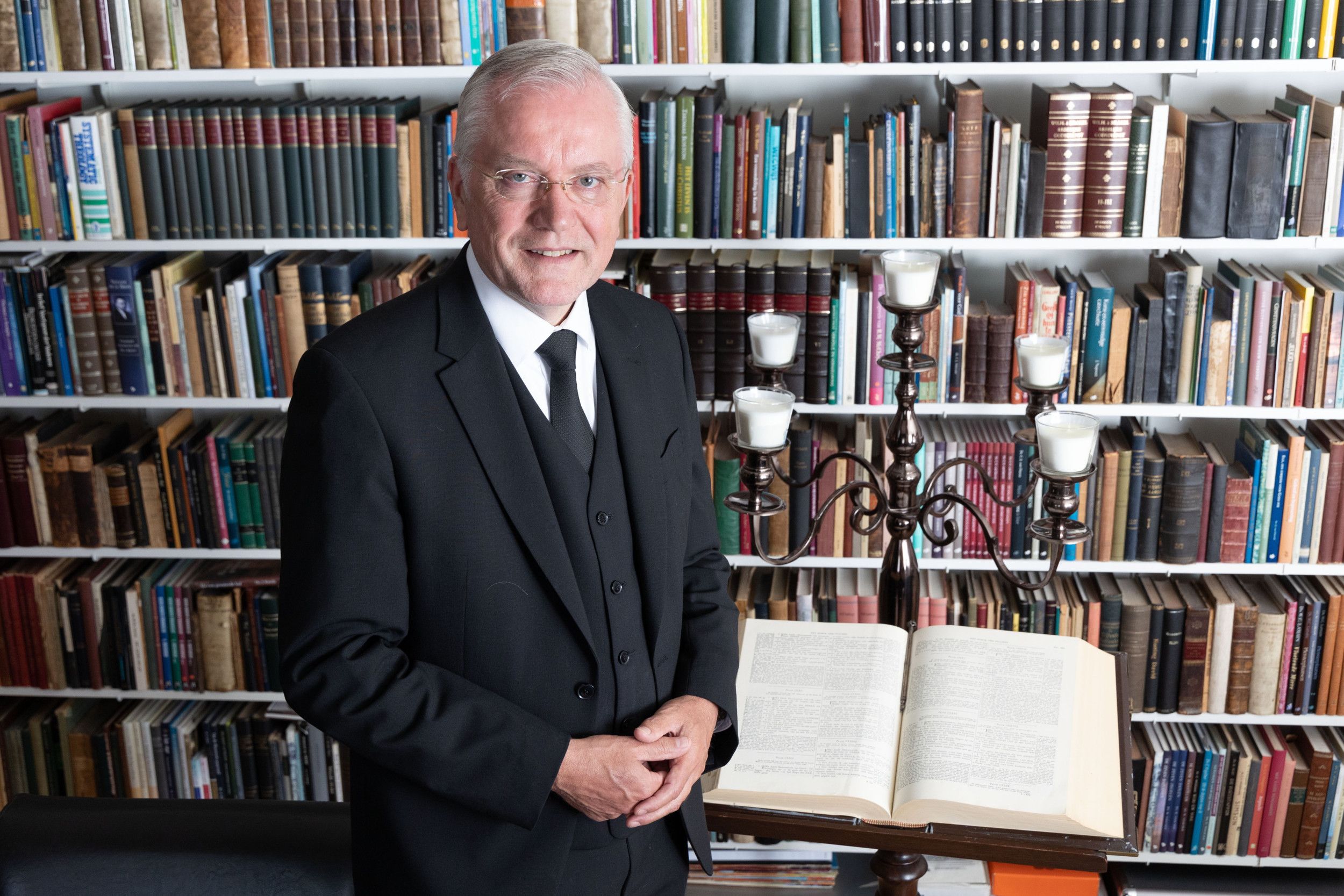 Man met bril en zwart pak voor een boekenkast.