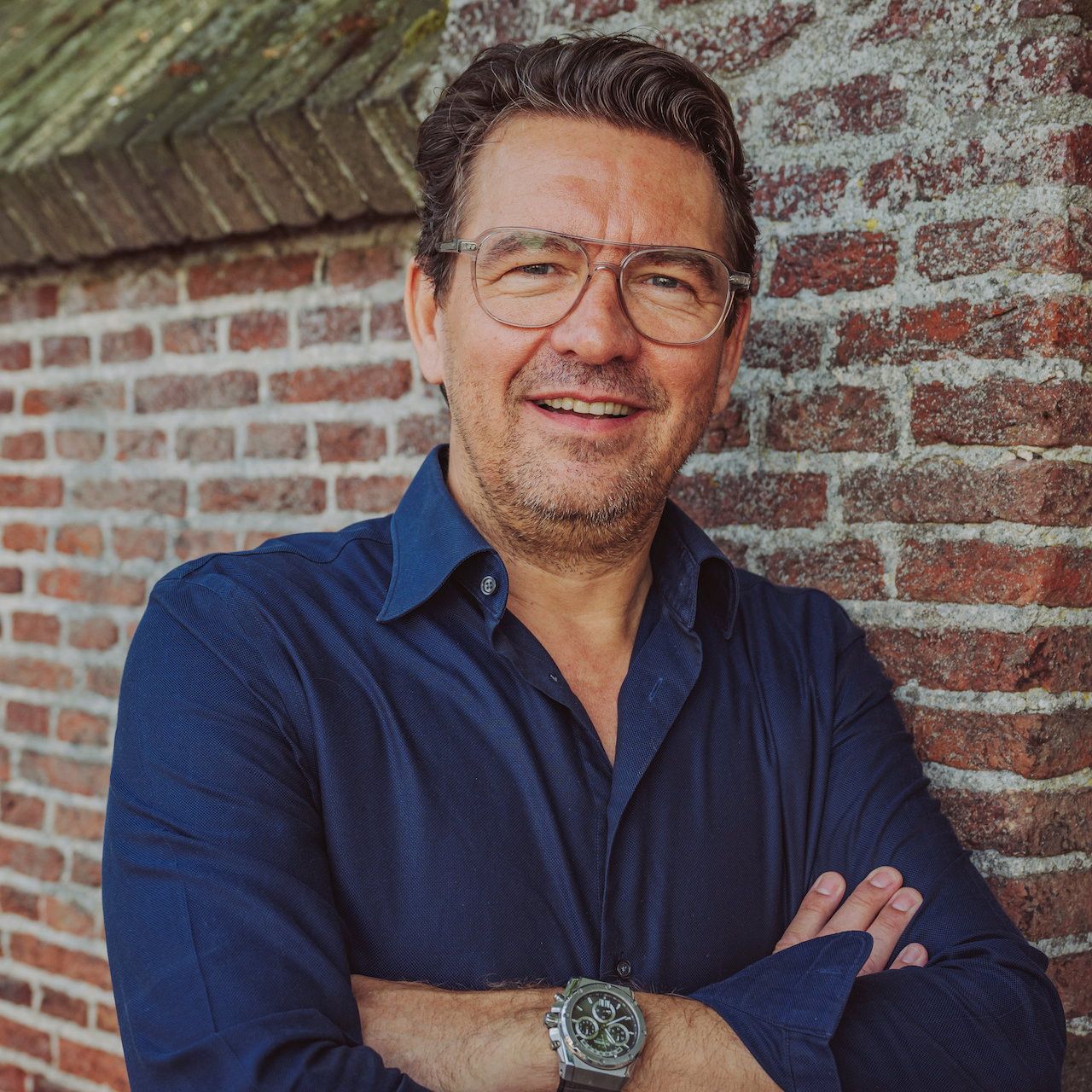 Man met bril en blauw overhemd voor een muur.