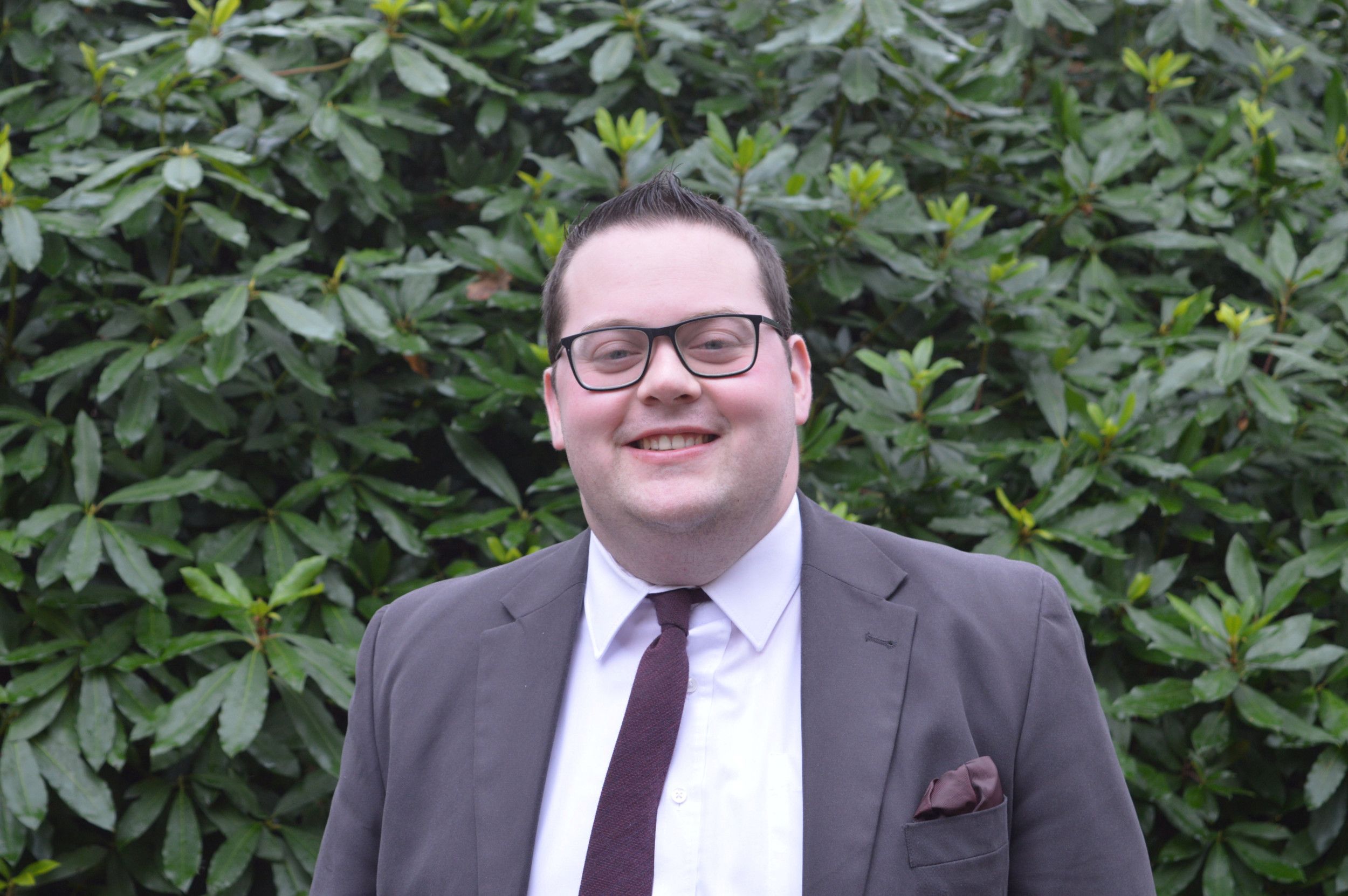 Man met donkere bril en grijs kostuum voor een groene haag.