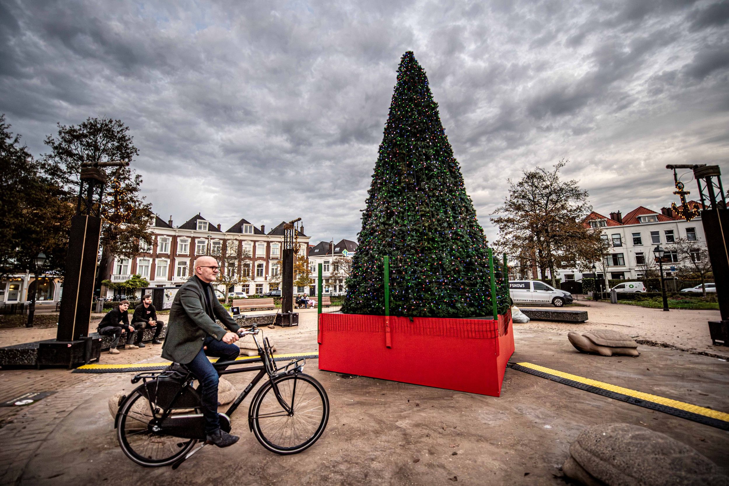 Man op een ouderwetse fiets naast een oranje bak met daarin een kerstboom vol lichtjes.