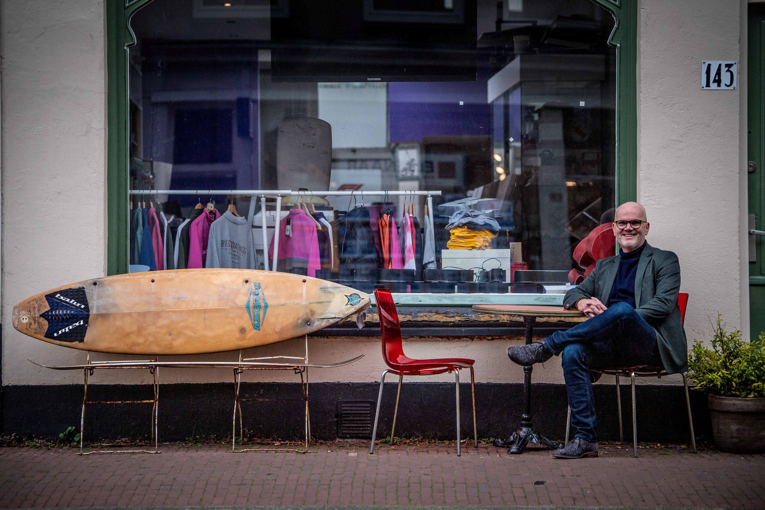 Donker geklede man, zittend op een stoel voor de etalage van een winkel met een roe vol kleurige truitjes.