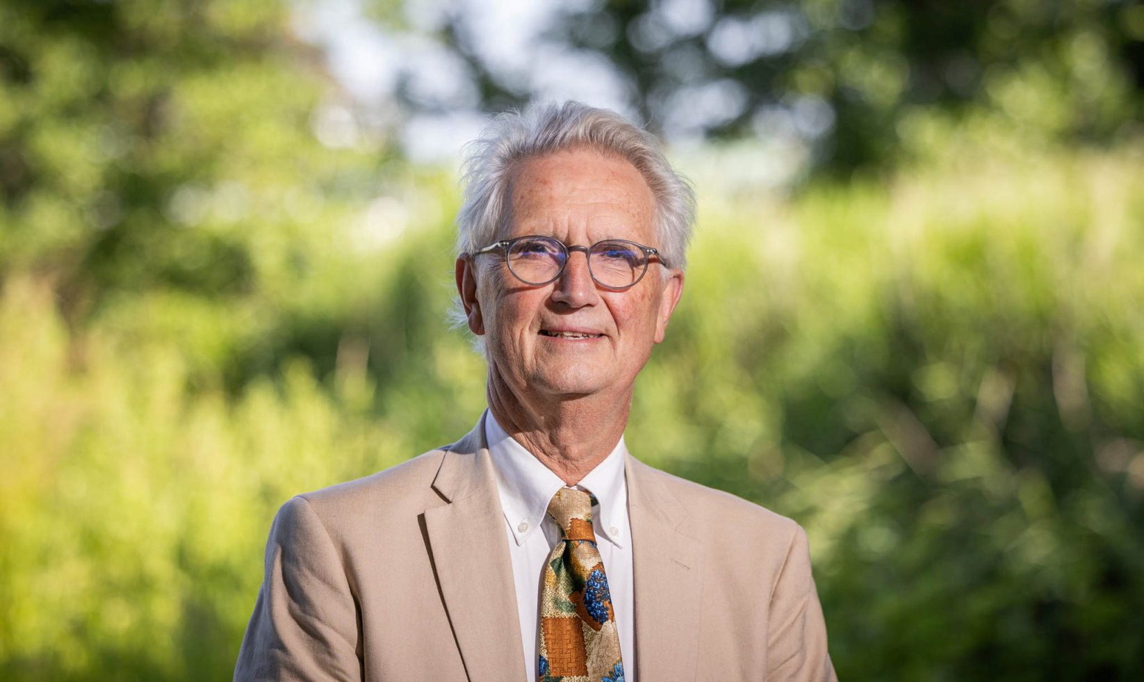 Man met bril en grijs haar draagt een beige jasje en veelkleurige stropdas. 