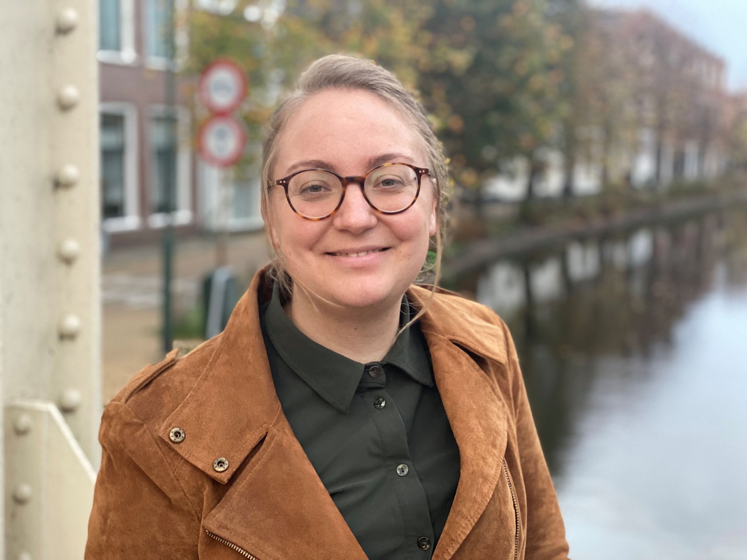 Vrouw, dertiger, blond haar en bril met bruin jasje aan en donkergroene blouse.