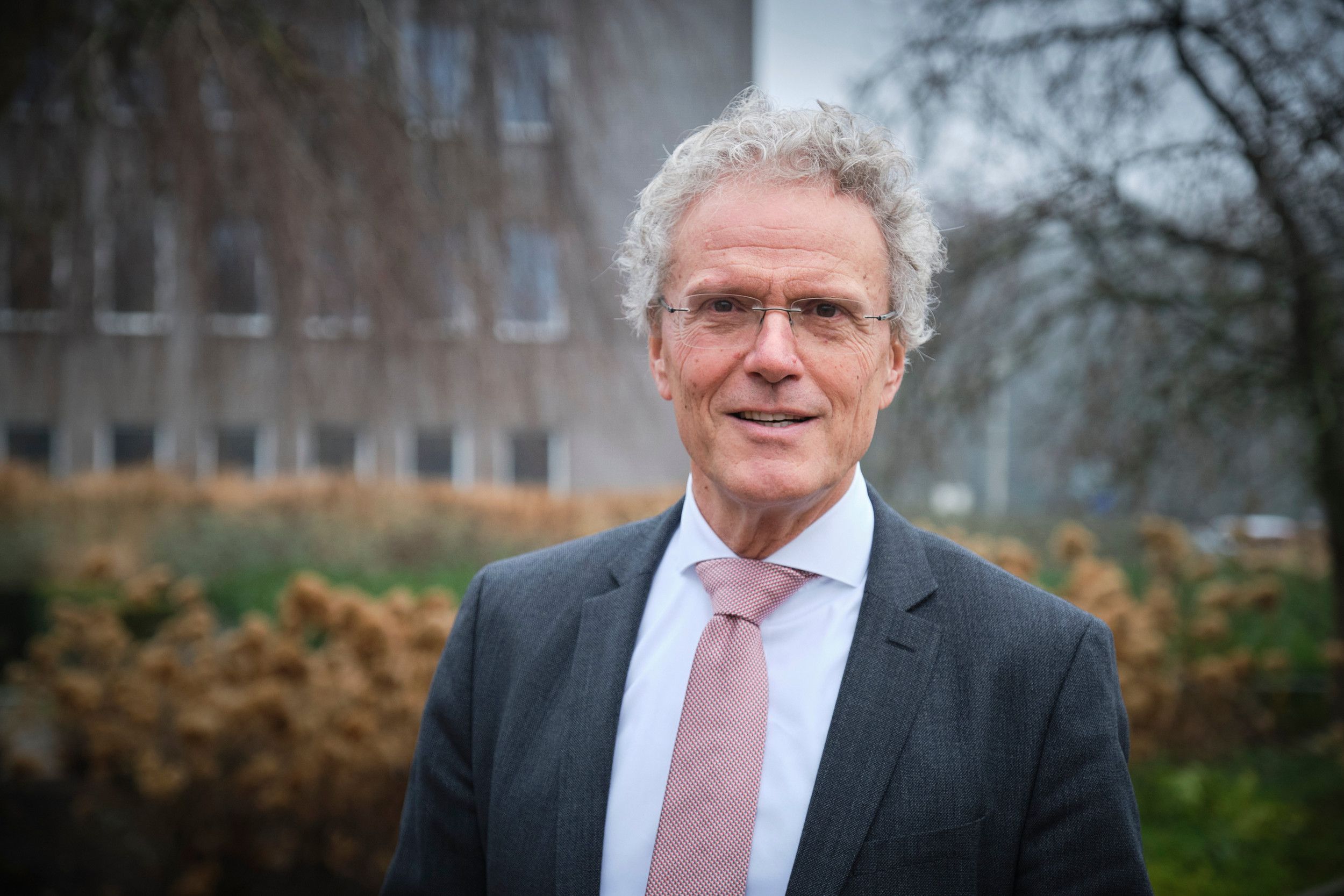 Portret van man met grijs haar en bril, gekleed in een grijs pak met overhemd en stropdas.