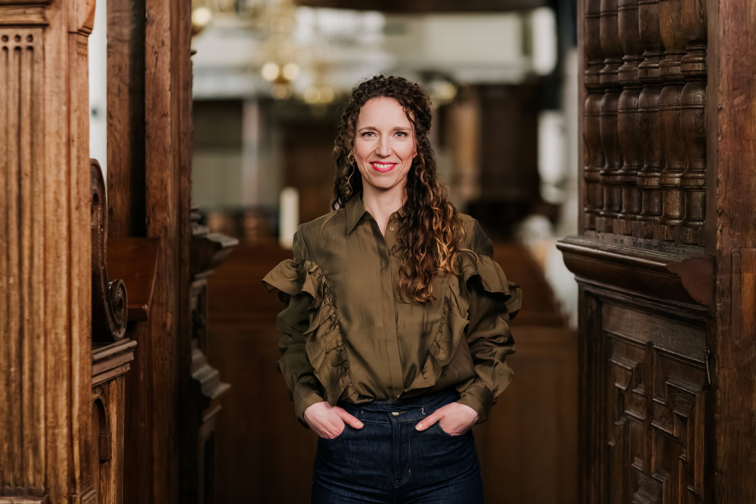 Vrouw met lang, krullend haar, gekleed in een spijkerbroek en blouse, met de handen in de zakken.