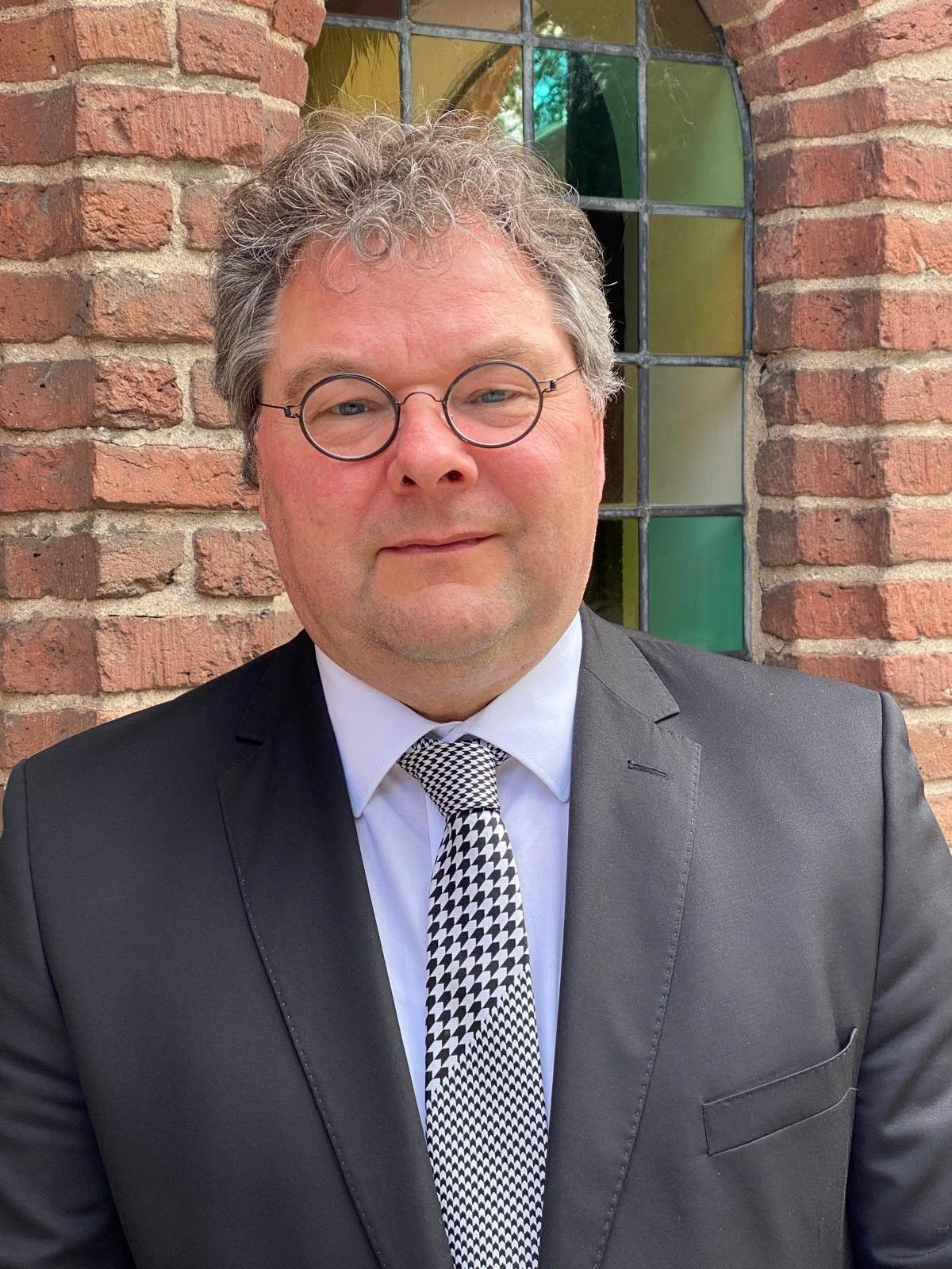 Man met grijzend haar en bril in donker kostuum.