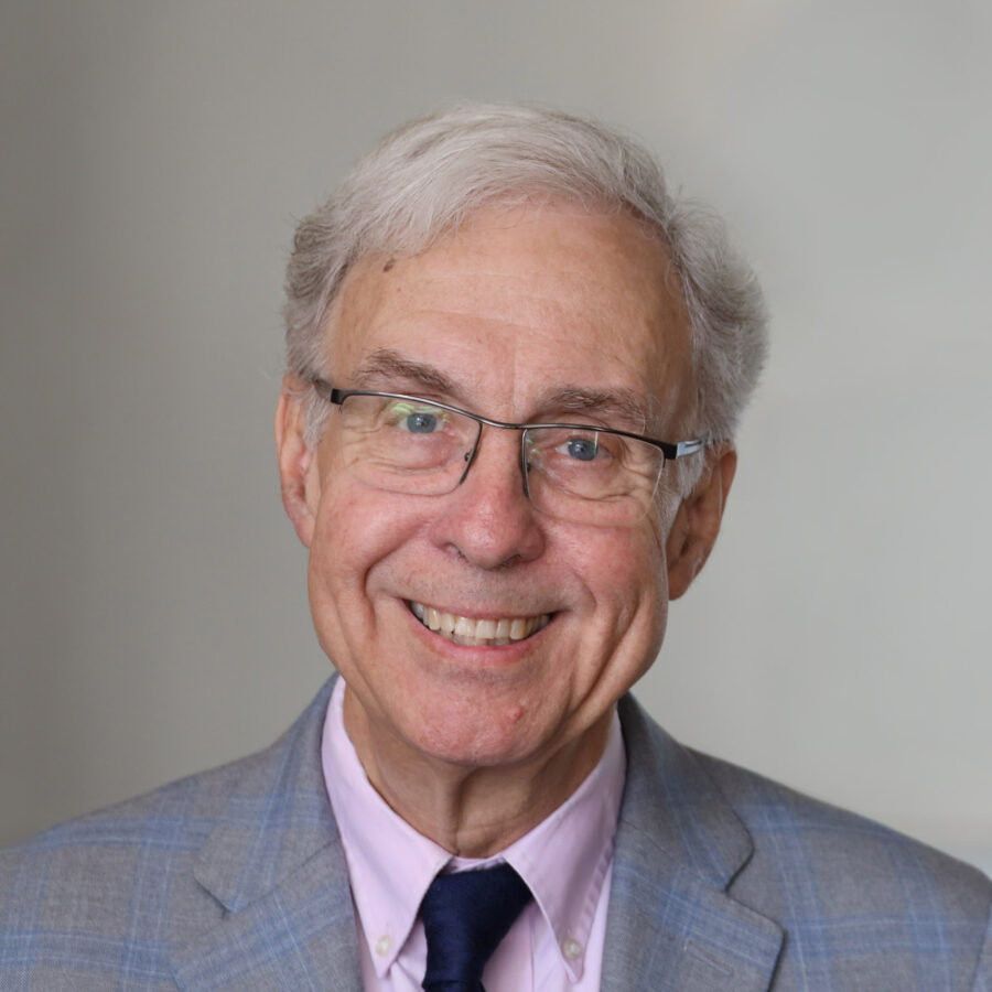 Oudere man, grijs haar, met bril op. Hij draagt een roze overhemd, een donkerblauwe stropdas en een lichtgeruit colbert.