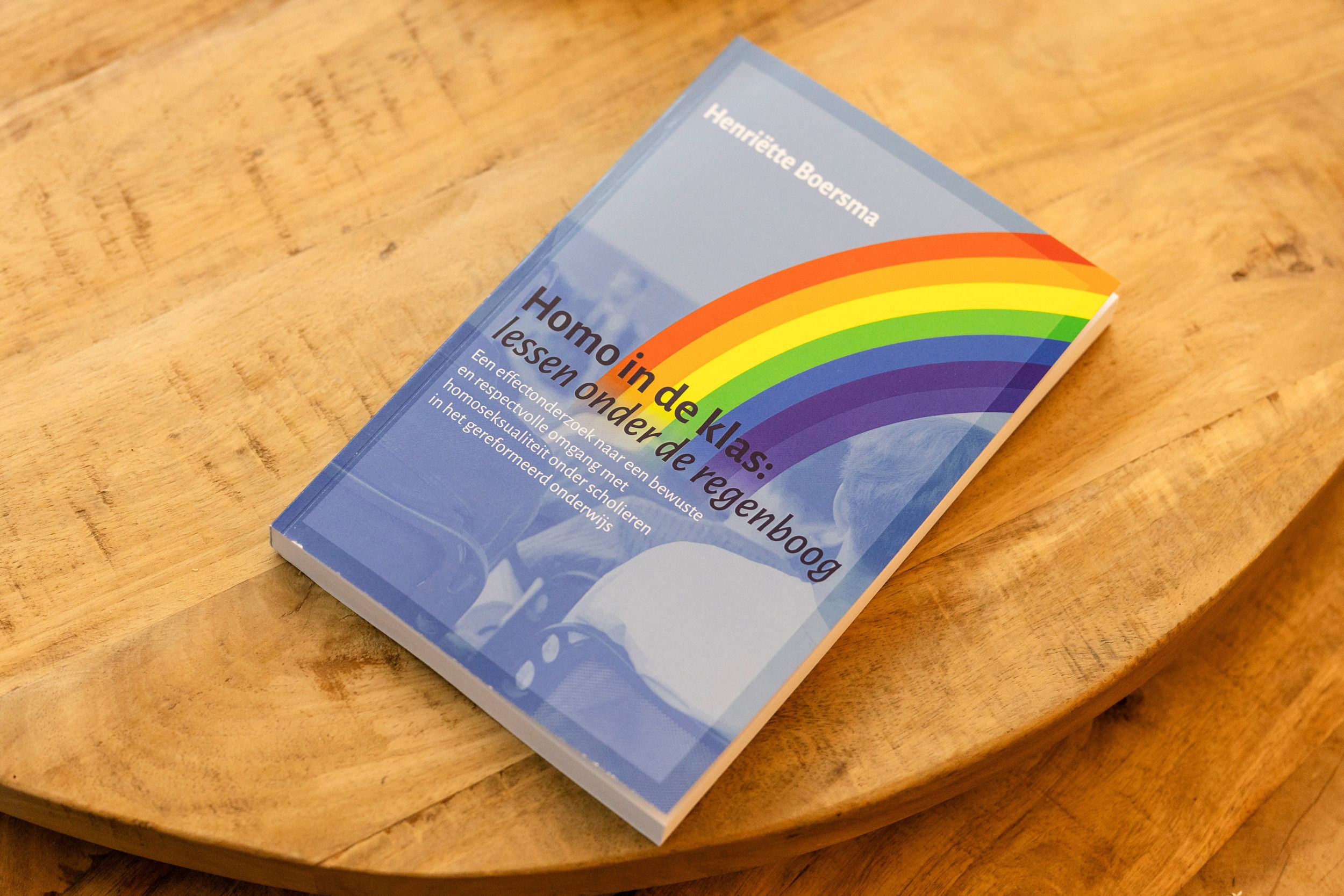Foto van een boek met blauwe kaft en regenboog en zittende jongeren, van achteren gezien.