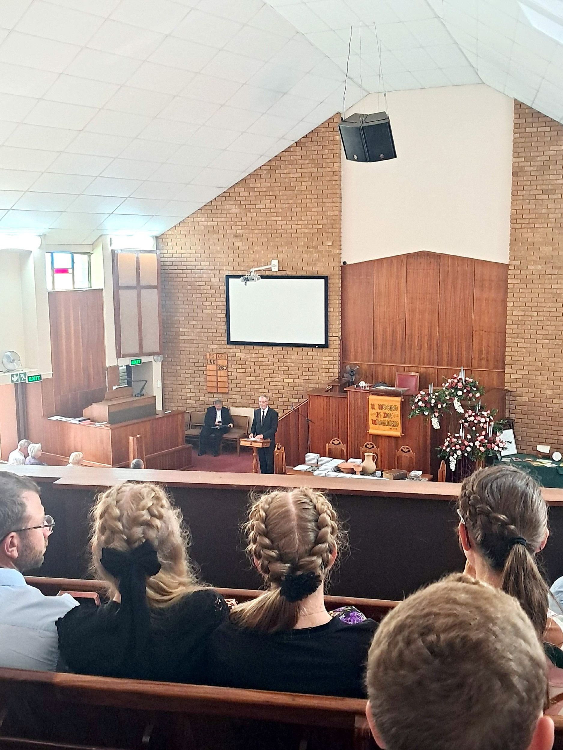 Galerij in een kerk waar meisjes met gevlochten haren zitten. In de kerk een kansel, met daarvoor een liturgische tafel waarop een Avondmaalsstel staat en boeken liggen. Achter de katheder staat een man in een zwart pak.