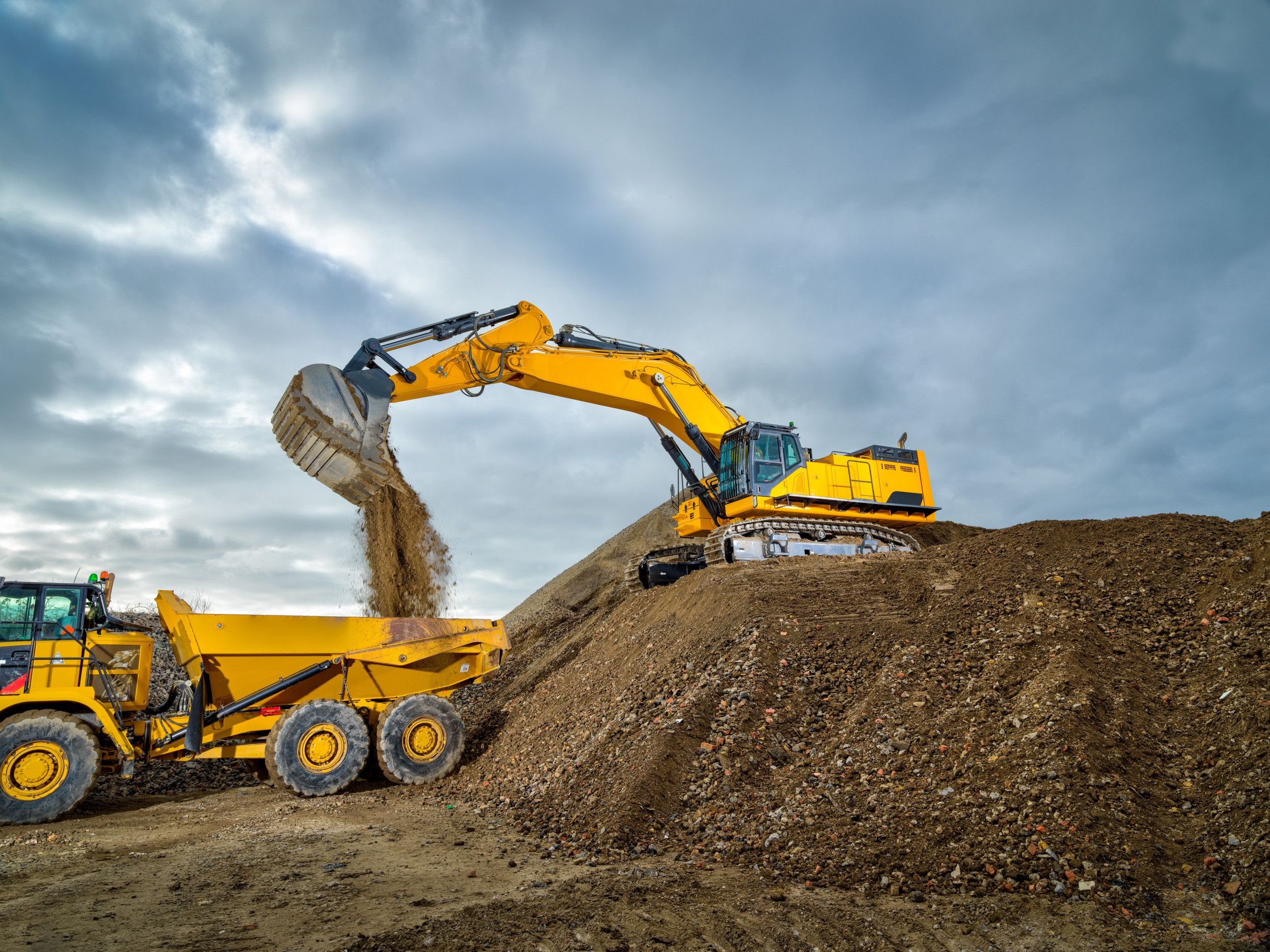 Foto van graafmachine die grind en zand schept in een dumptruck op een bouwplaats.