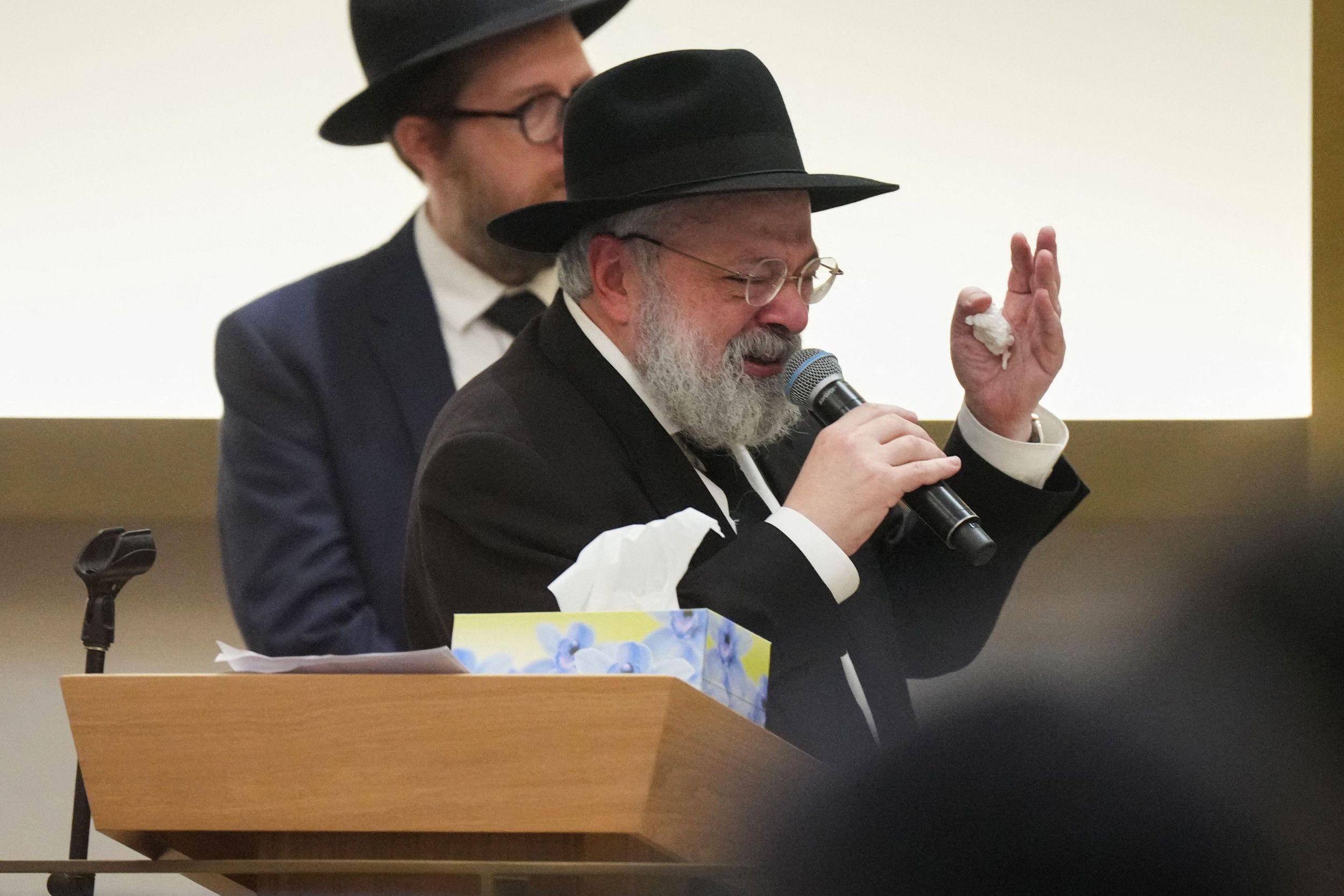 Man in zwart pak met zwarte hoed op spreekt in microfoon.