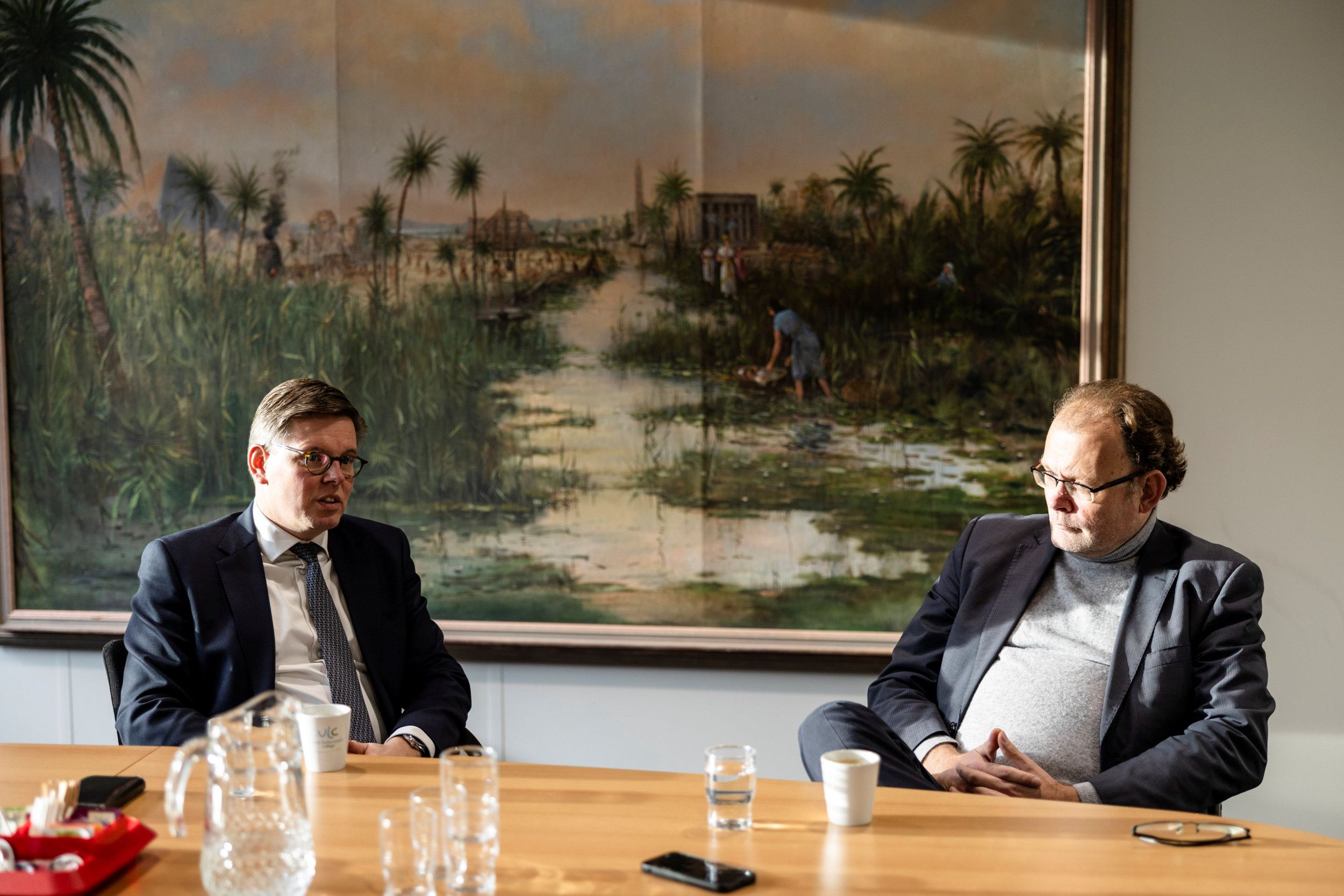 Twee mannen aan een tafel. Op de achtergrond een schilderij met het Bijbelse tafereel waarbij Mozes in een biezen mandje ligt. 