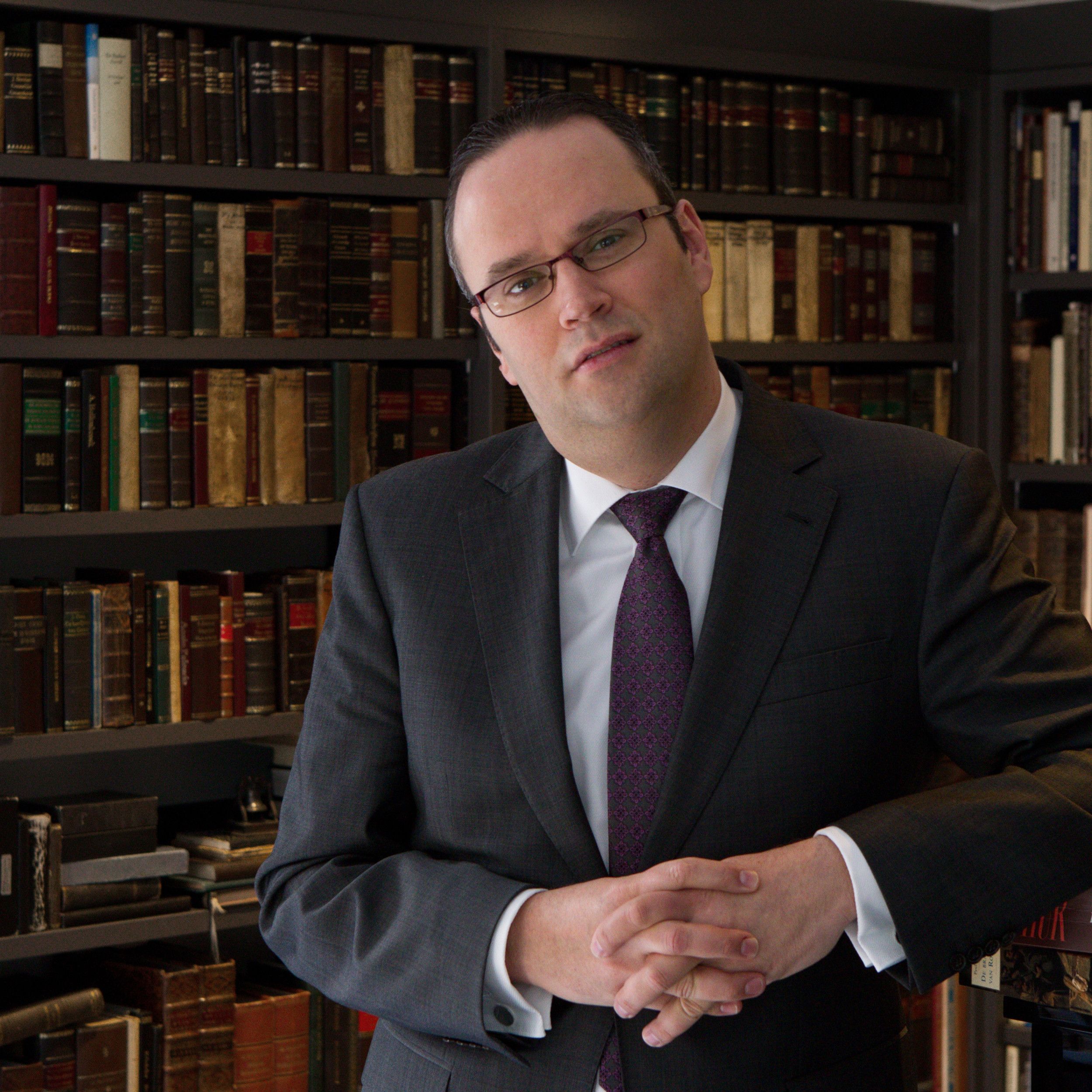 John Tollenaar, een man met bril, donker kostuum, voor een boekenkast.