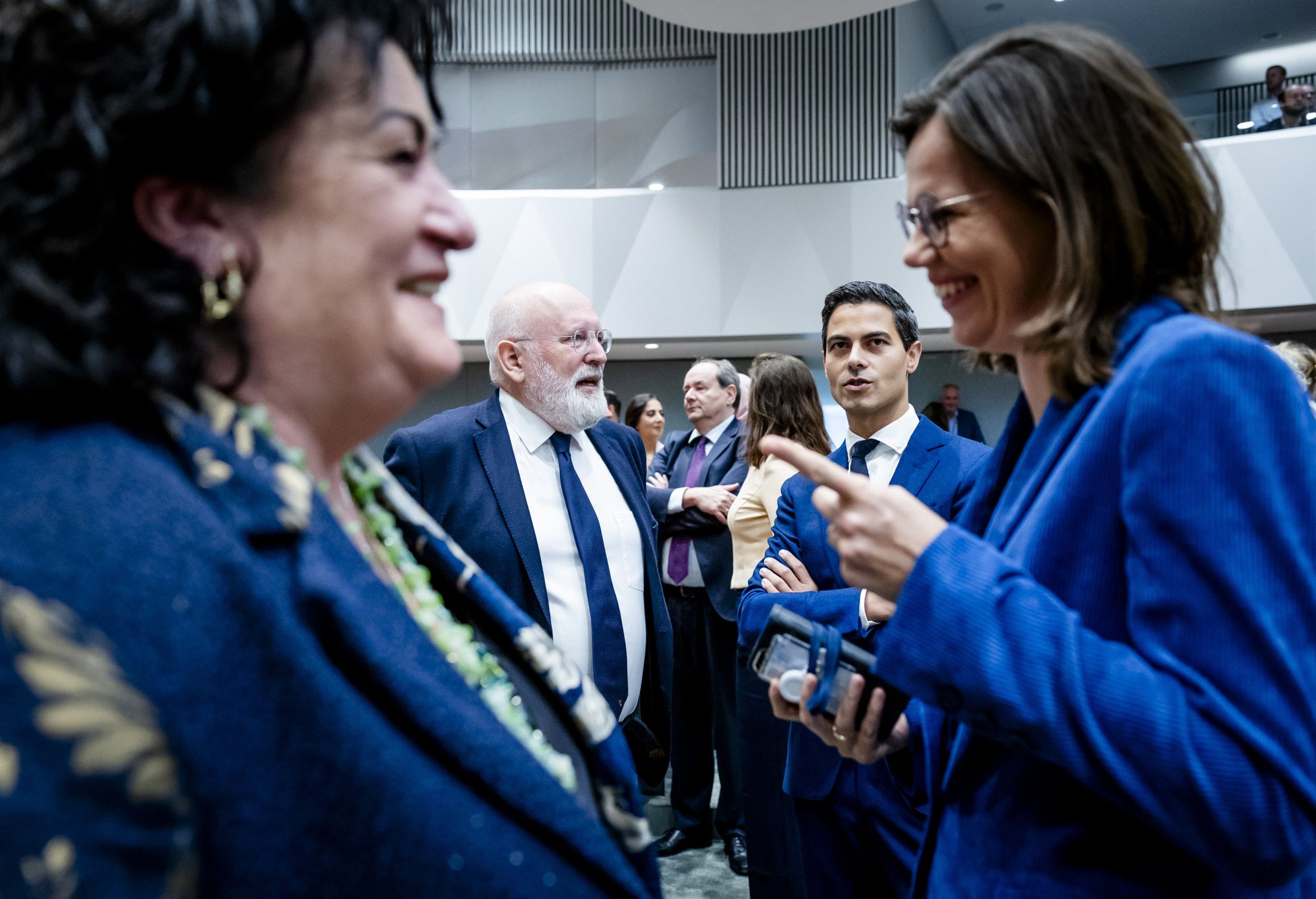 Twee vrouwen, allebei in helderblauwe kleding, lachen naar elkaar in de plenaire zaal van de Tweede Kamer. Naast hen staan andere Kamerleden te praten.