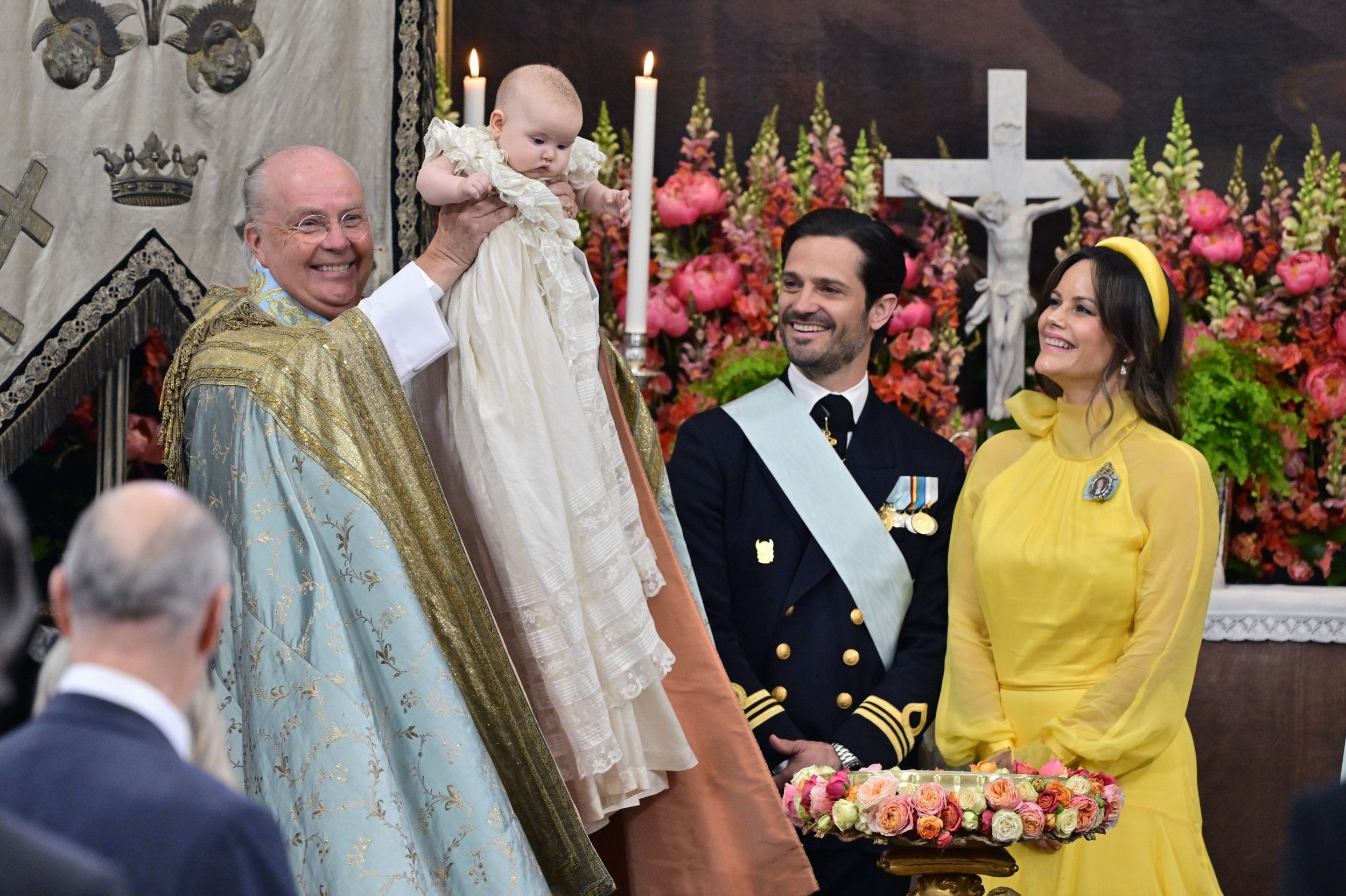 Lachende man in bisschopsmantel houdt baby omhoog. Man in uniform en vrouw in gele jurk kijken toe.