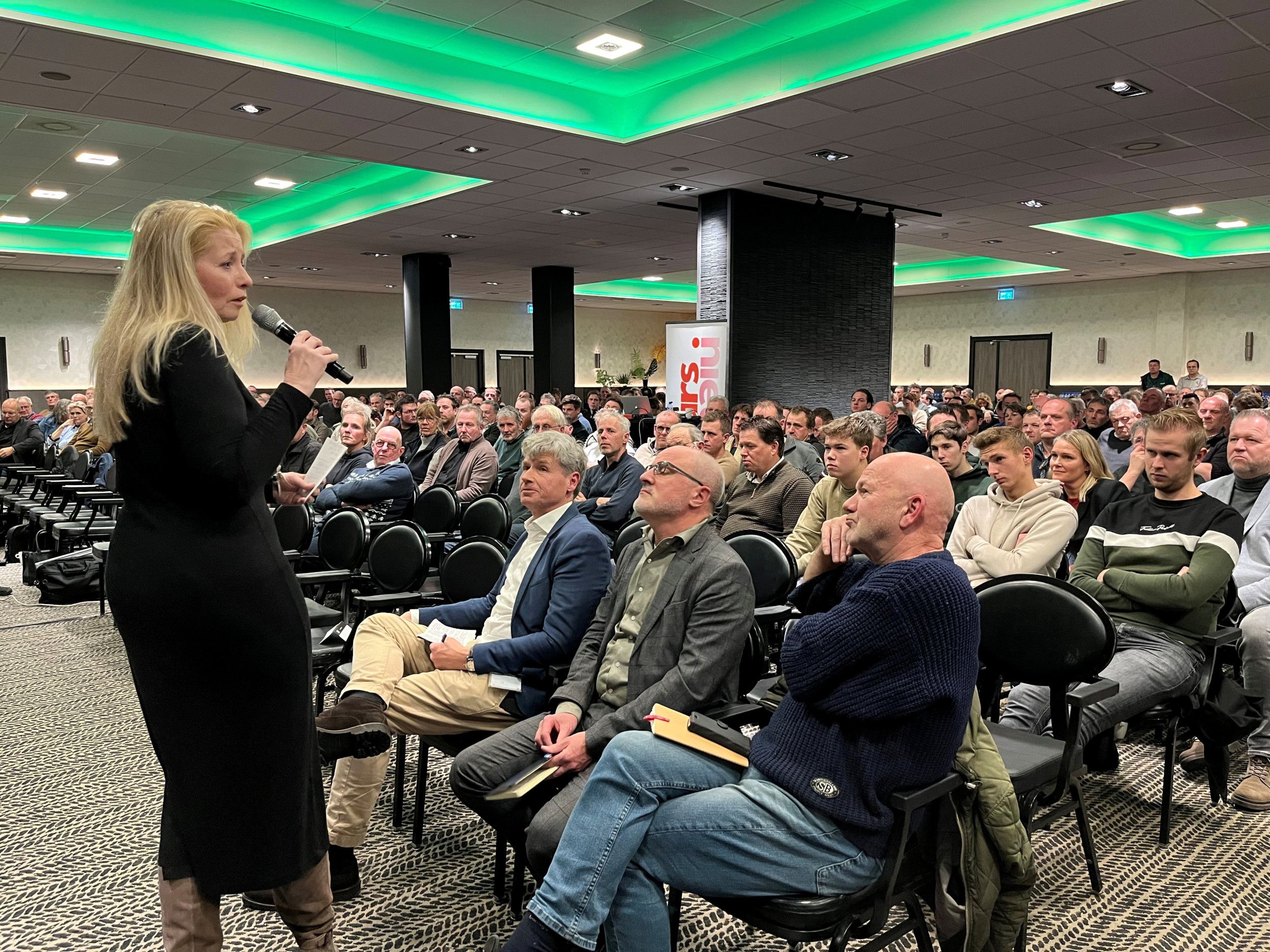 Een blonde vrouw met een microfoon in de hand staat voorin een zaal met overwegend mannen.
