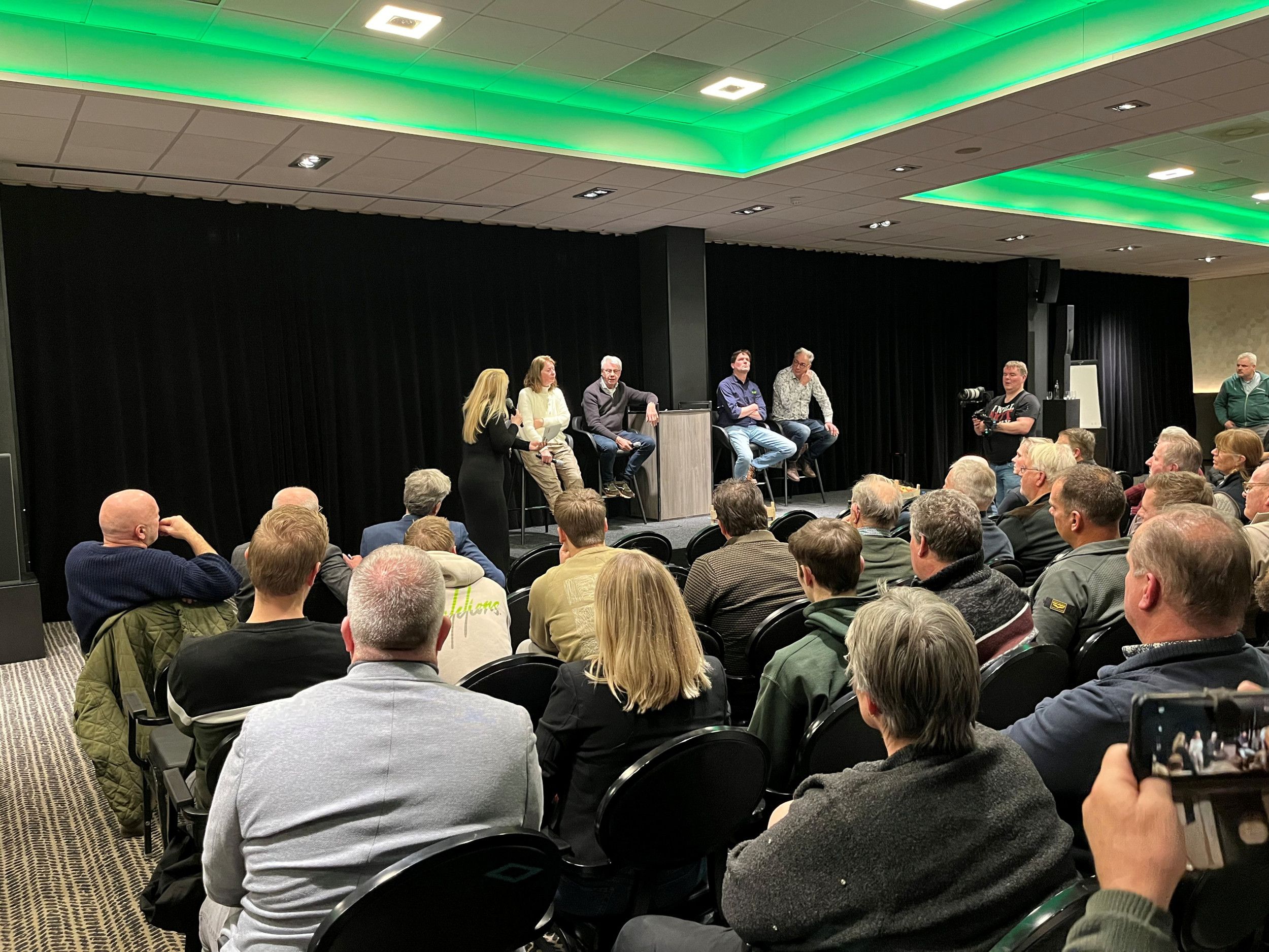 Een vrouw en drie mannen zitten op krukken op een podium in een zaal vol mensen.