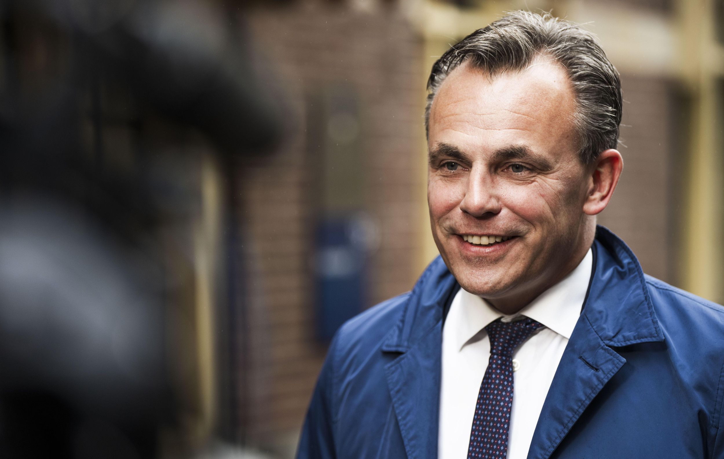 Glimlachende man in een blauwe jas. Hij draagt een donkere stropdas op een wit overhemd.