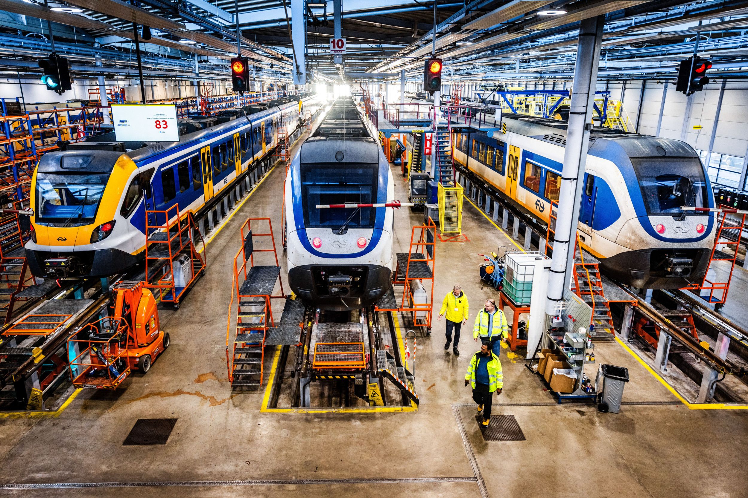 Drie treinen staan op verhoogde sporen in een bedrijfshal. Ervoor lopen mannen in lichtgevende jassen.