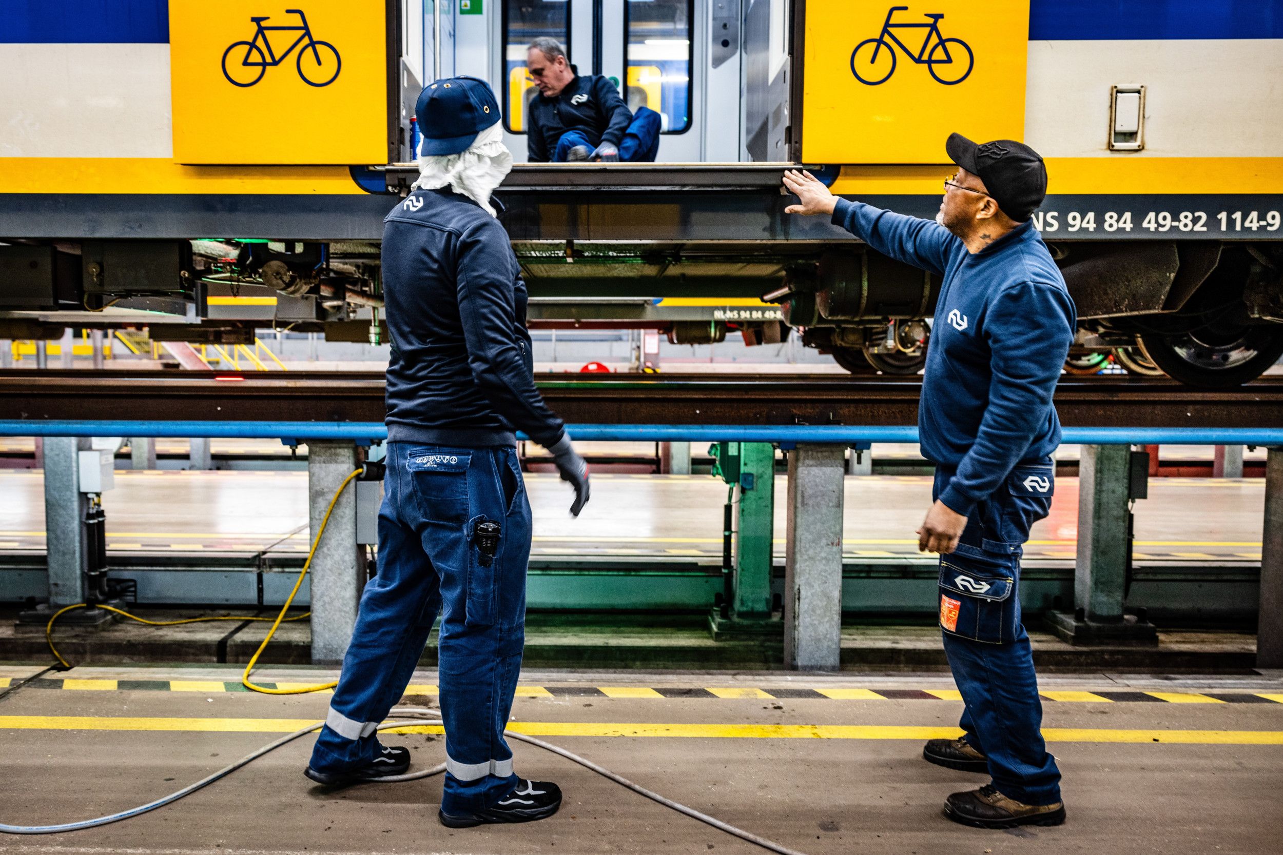 Twee mannen in donkerblauwe werkkleding wijzen naar de openstaande gele schuifdeuren van een trein.