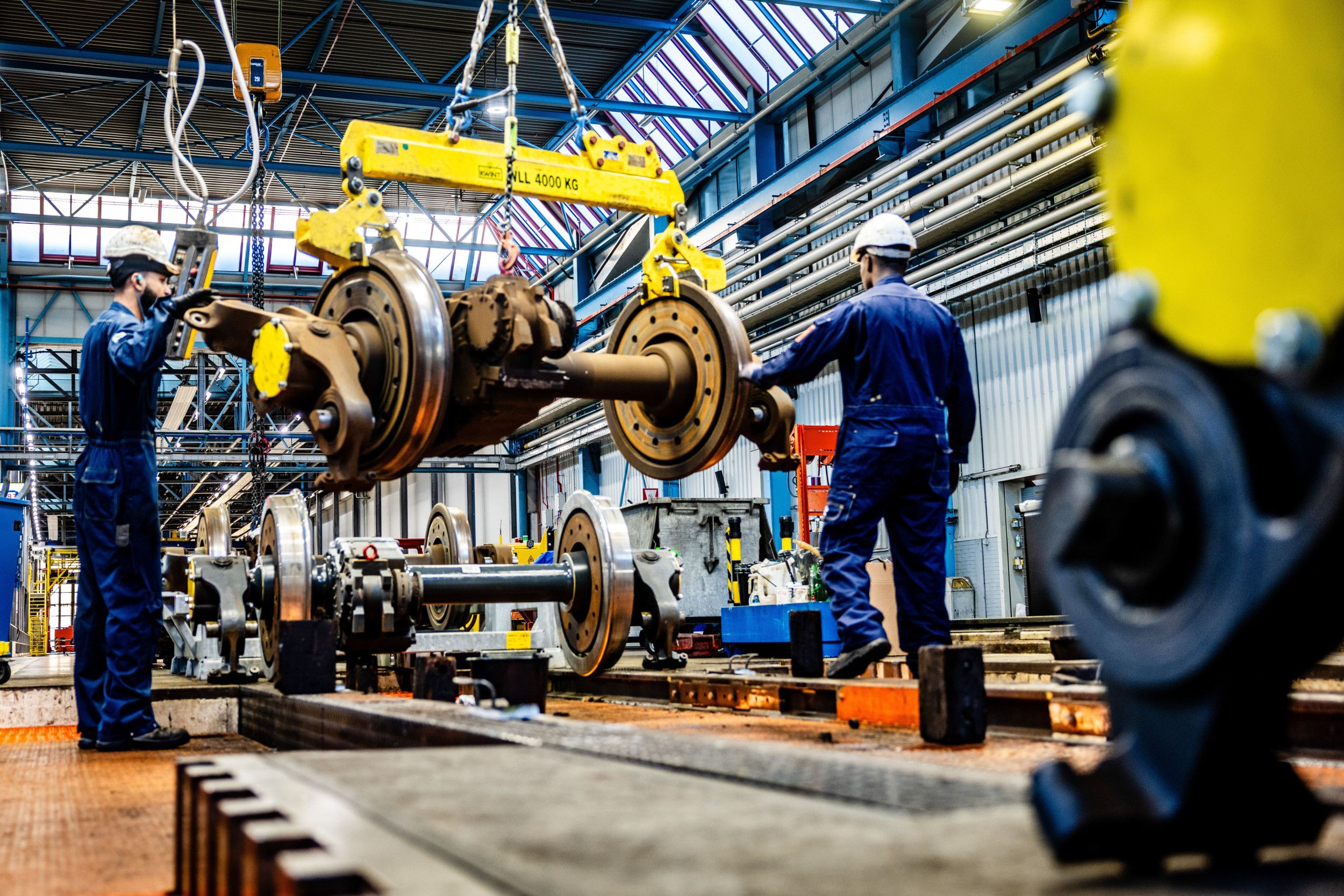 Twee mannen in blauw ketelpak en een witte helm op het hoofd verplaatsen met een takelsysteem een stalen treinwielset.