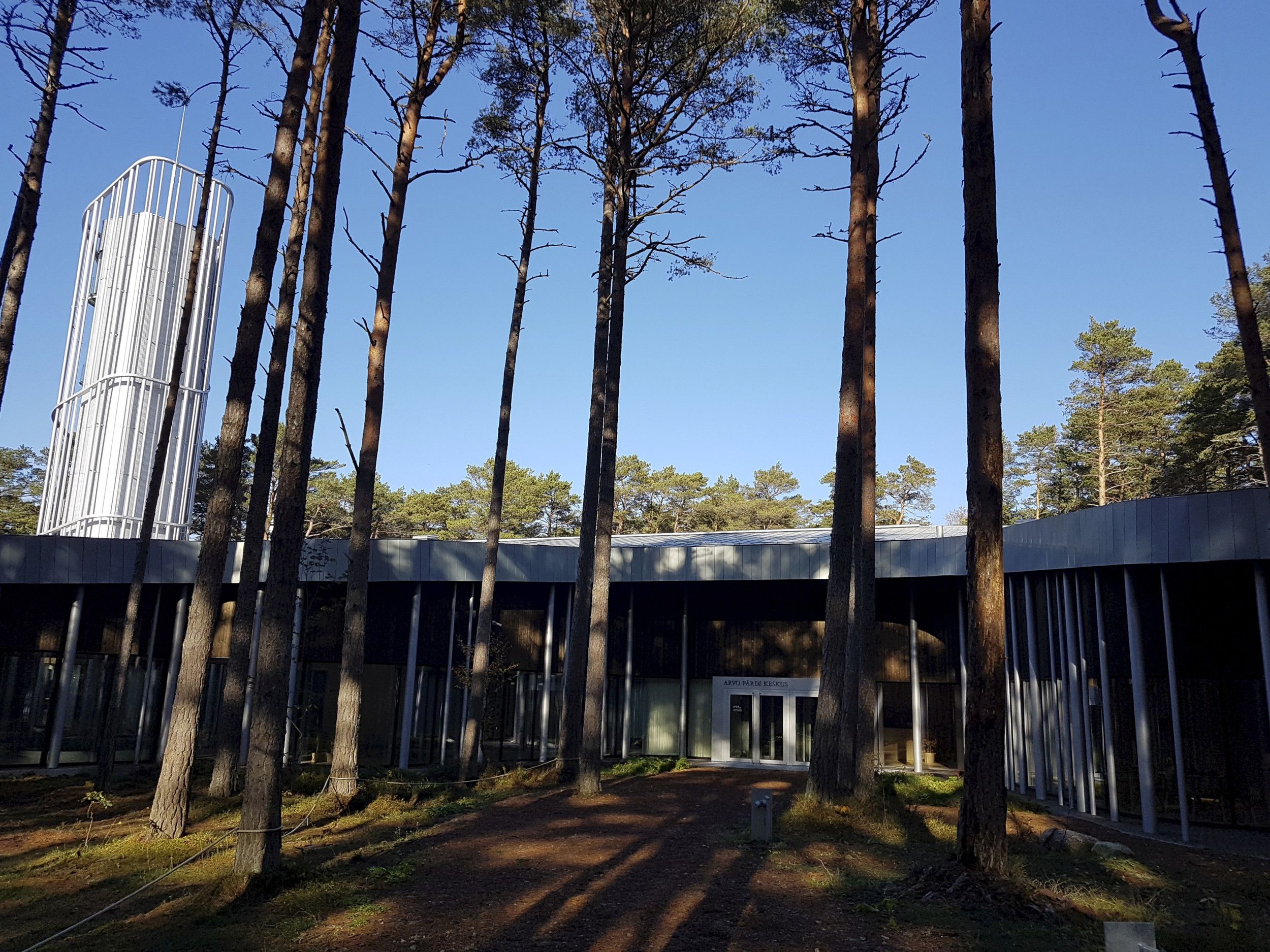 Gebouw in de bossen, met hoge bomen voor de ingang. Het is wit met zwart.