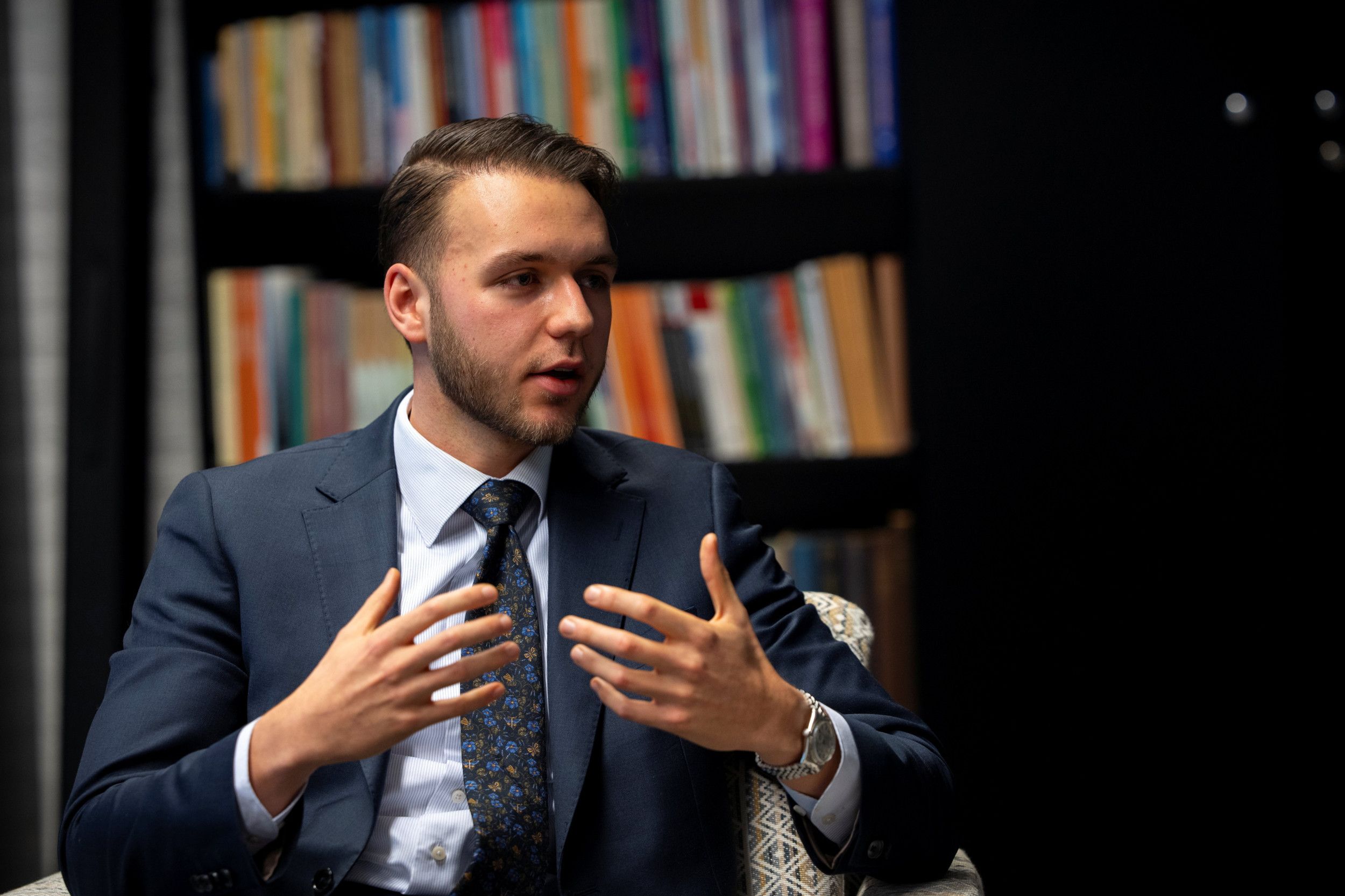 Jongeman met baardje, gekleed in een pak met stropdas, pratend en met zijn handen gebarend; hij zit op een stoel die voor een boekenkast staat.