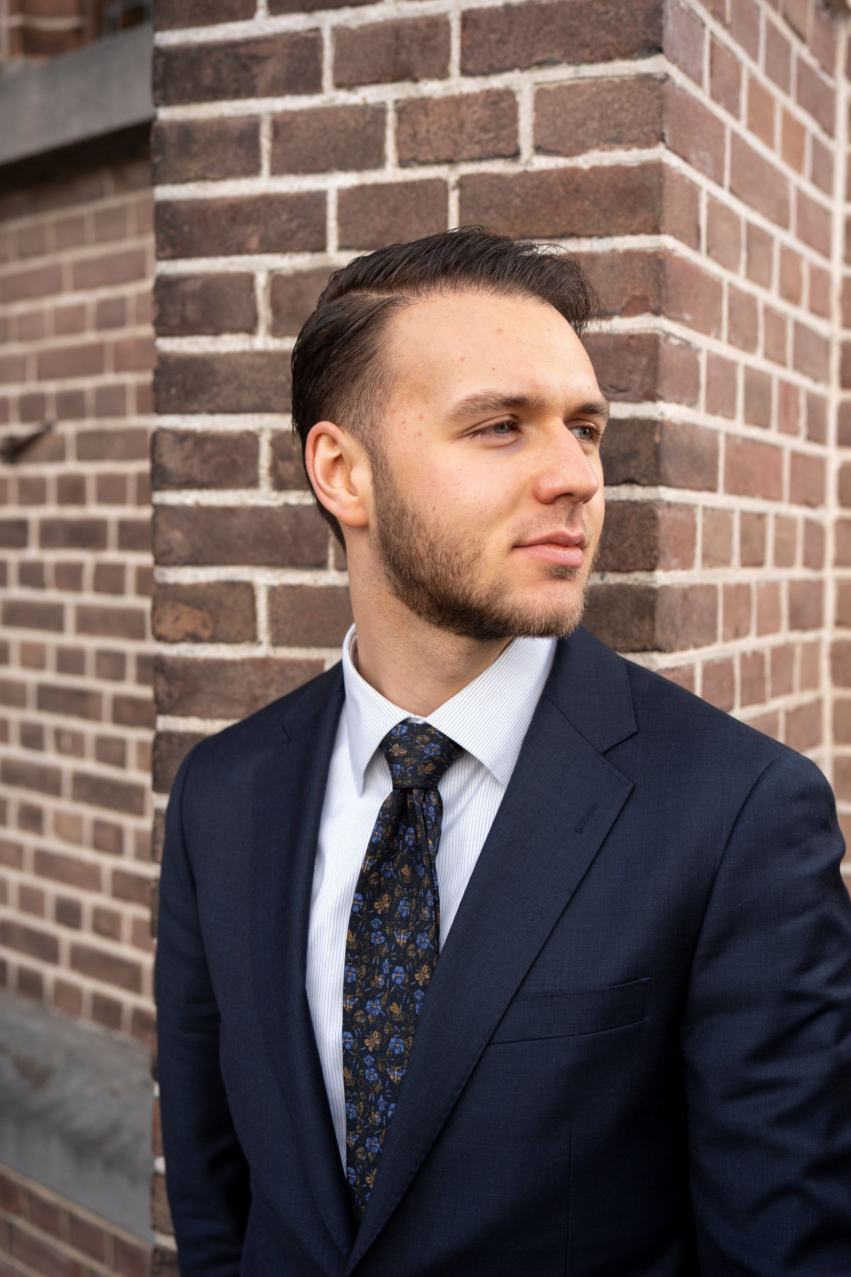 Jongeman met baardje, gekleed in een pak met stropdas, staat tegen de steunbeer van een oude kerk; hij kijkt naar rechts.