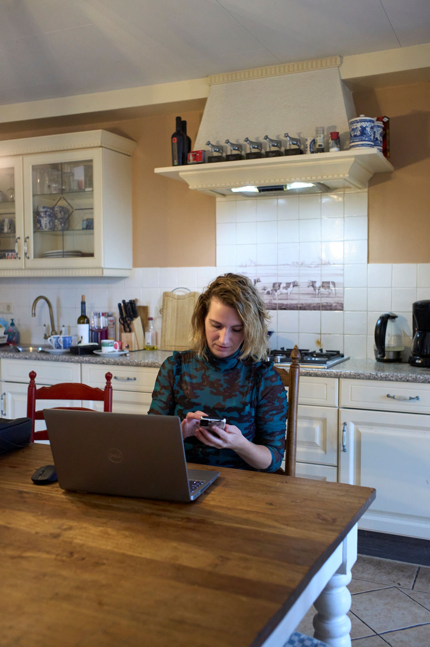 Jonge vrouw zit aan de keukentafel met een mobiele telefoon in haar hand.