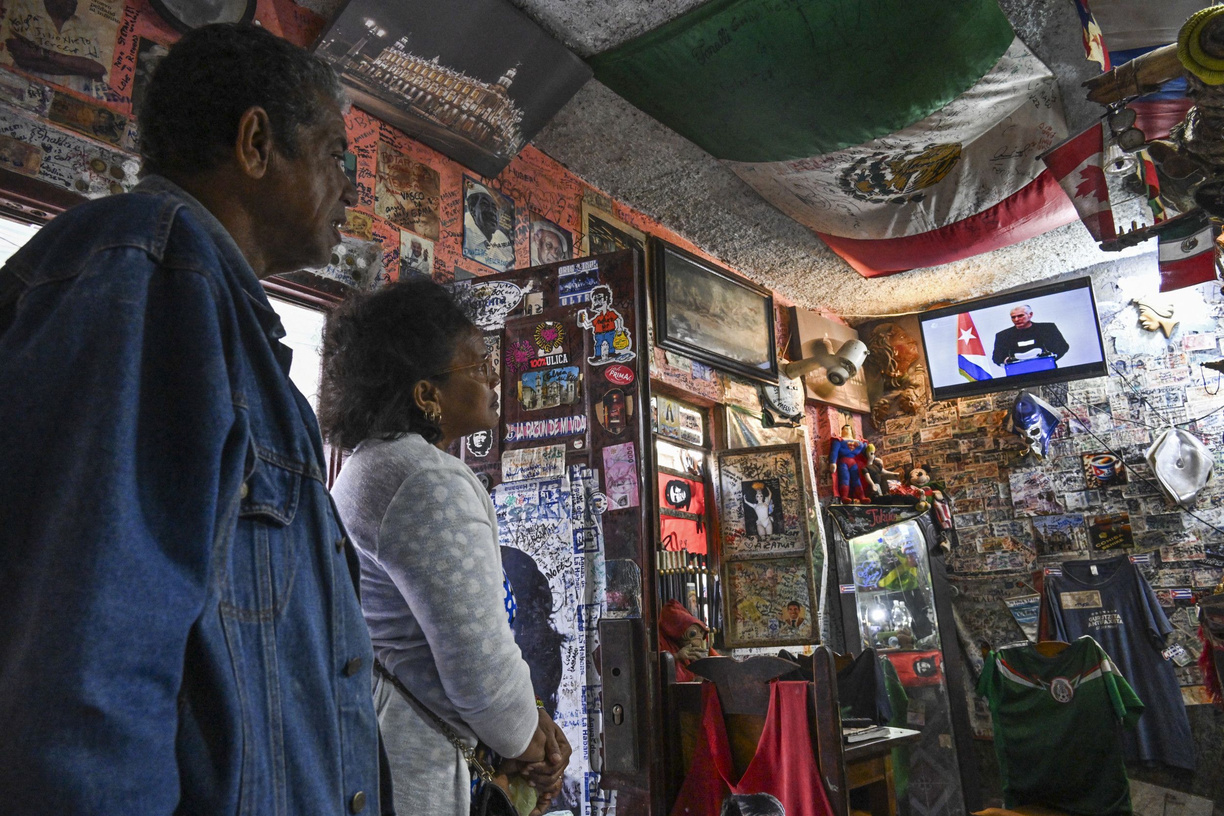 Een man en een vrouw kijken naar een beeldscherm met daarop een man achter een spreekgestoelte.