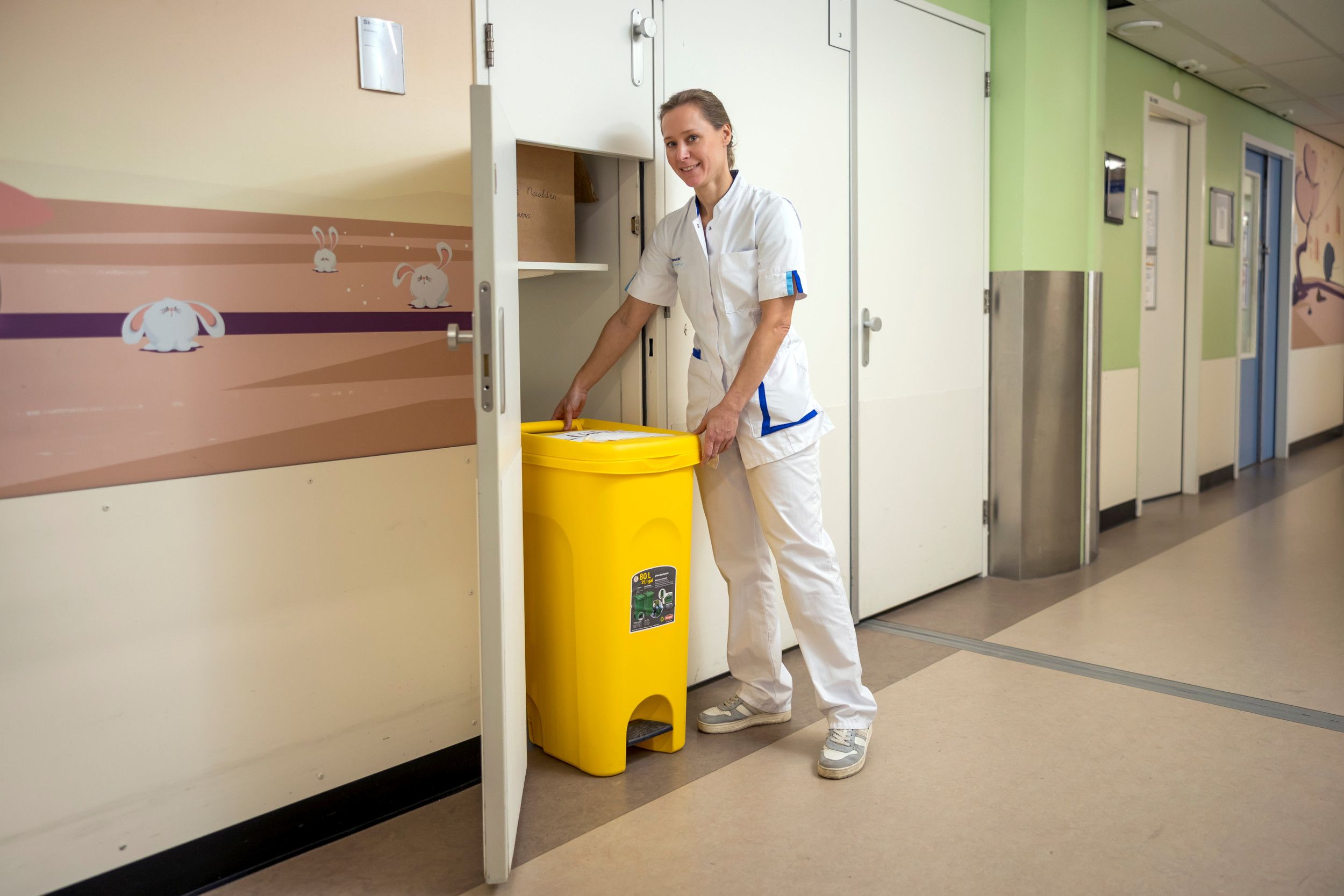 Een vrouw in witte jas bij een gele kliko in een gang van een ziekenhuis. 