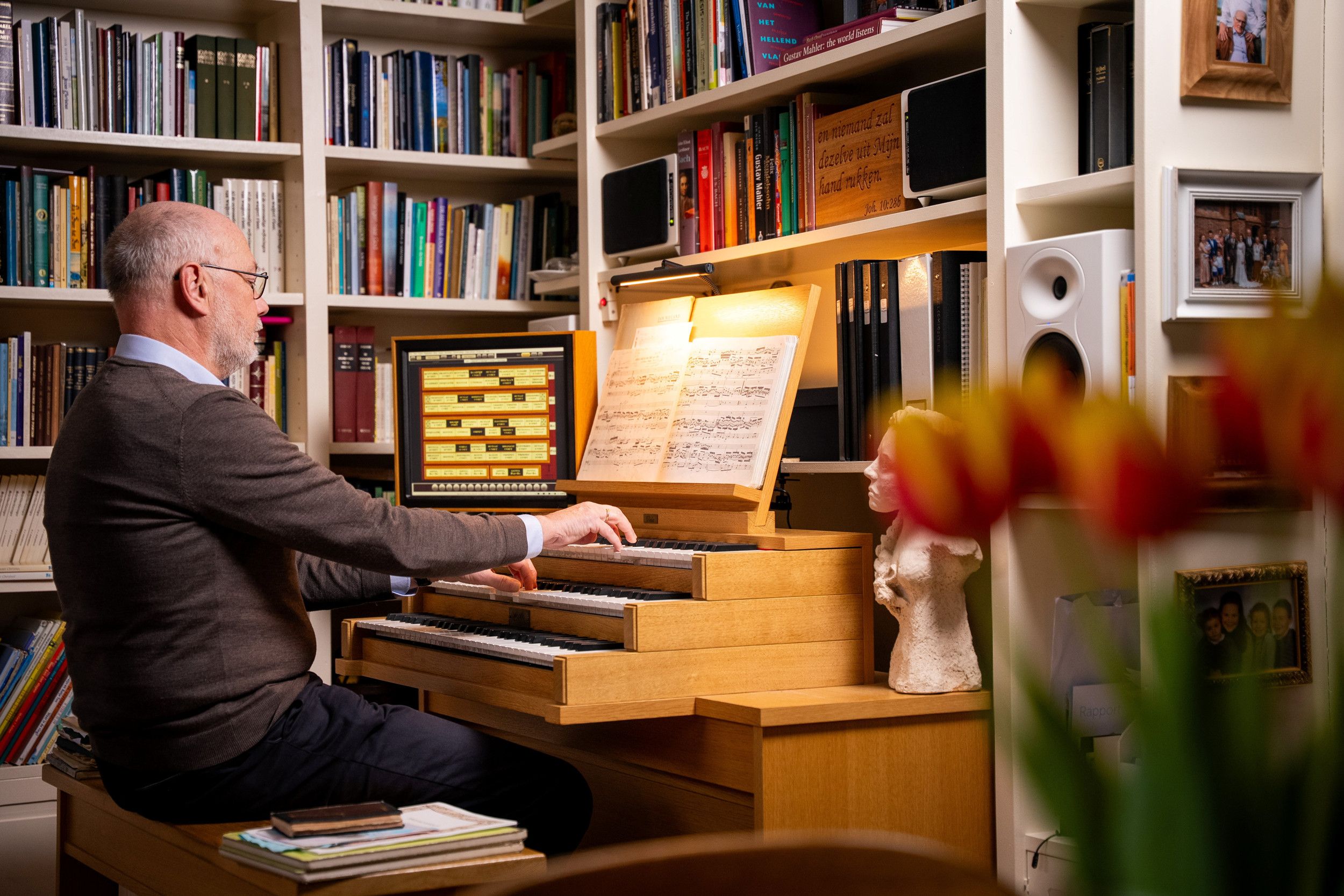 Een kalende man met bril bespeelt een elektronisch orgel dat in een woonkamer staat. Op de achtergrond zie je boekenkasten.