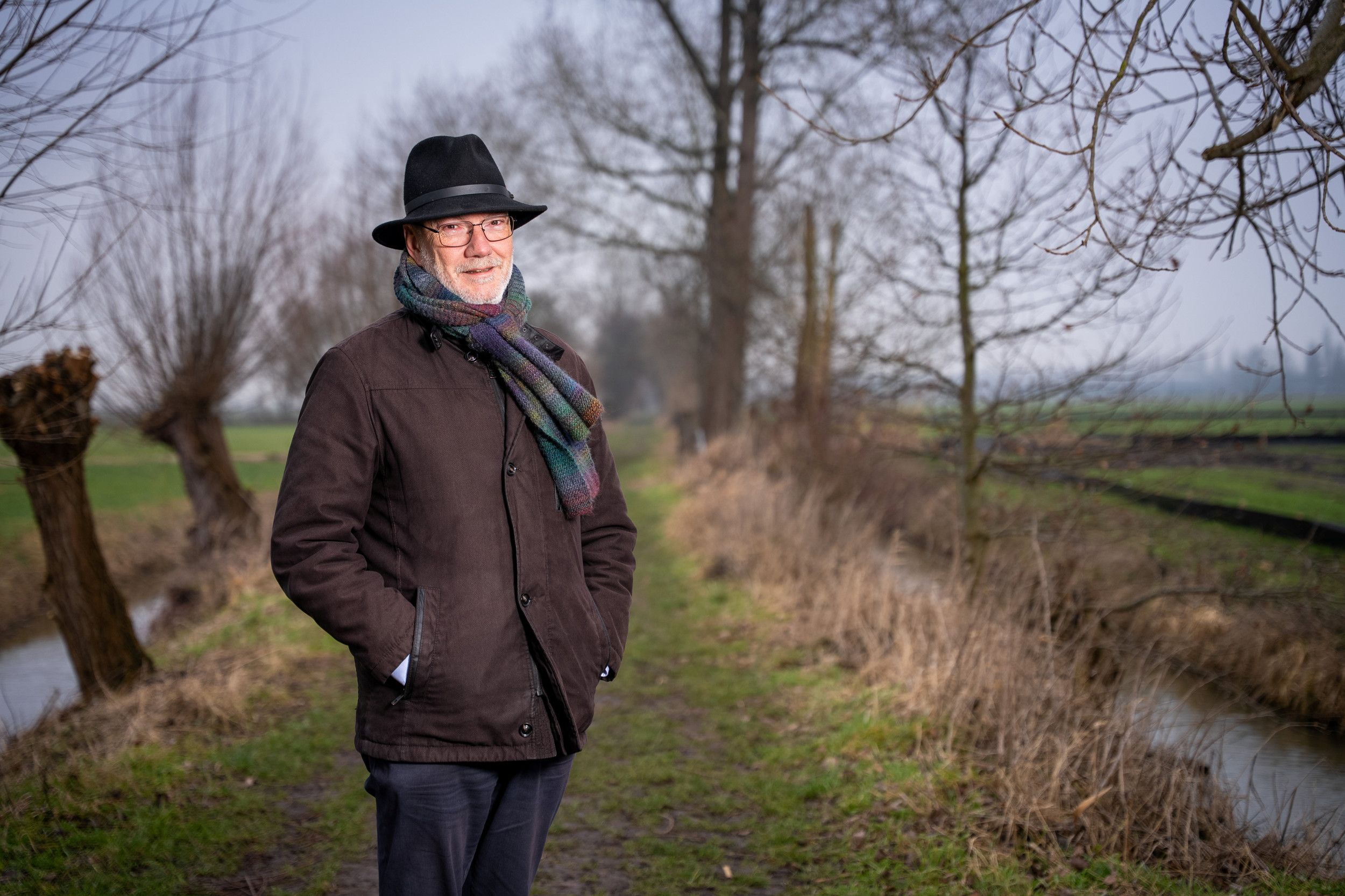 Een man in winterjas met sjaal en hoed staat op een landweg tussen twee sloten en weilanden.