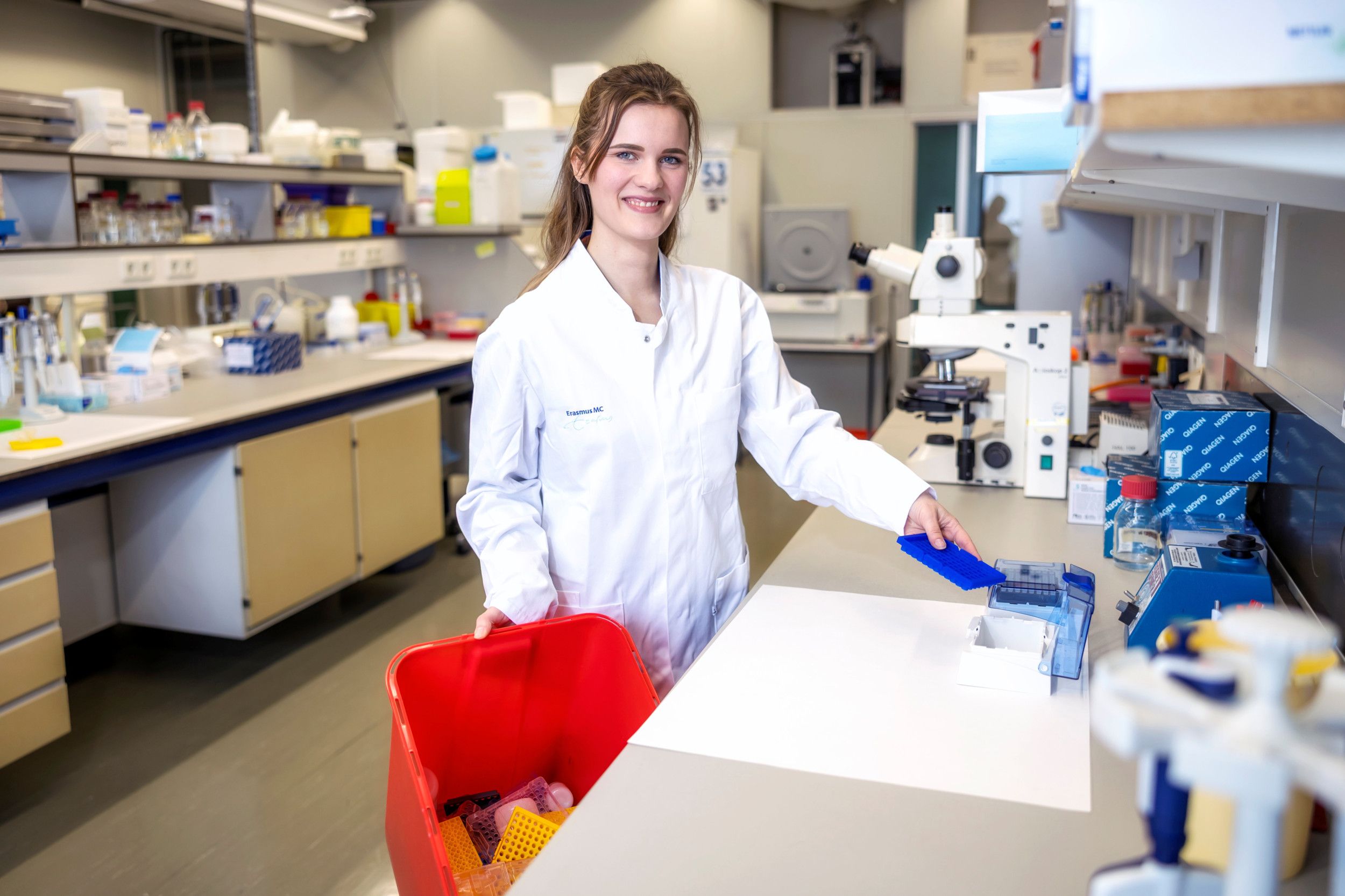 Een jonge vrouw in witte labjas staat in een lab. In haar ene hand heeft ze een houder voor pipetpuntjes, in haar andere hand een rode afvalemmer. 