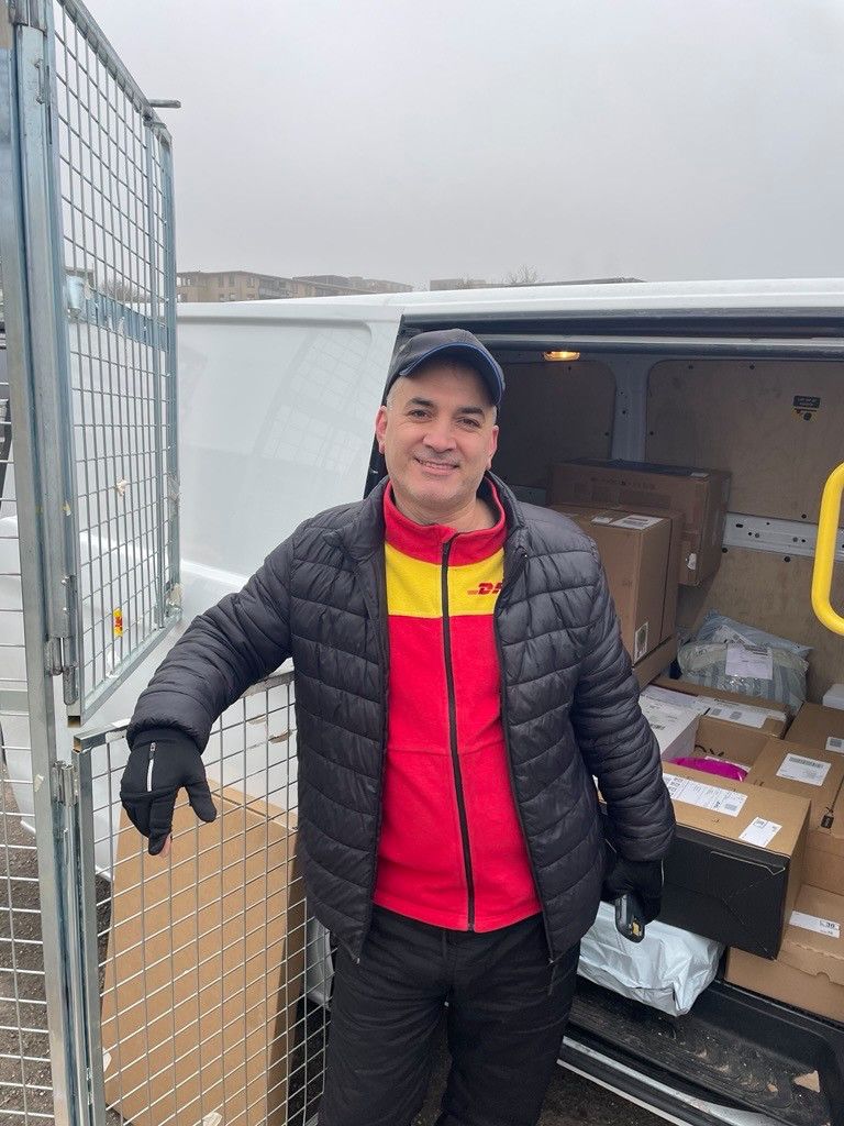 Een man staat voor zijn witte bestelbusje met een zwarte jas aan. 