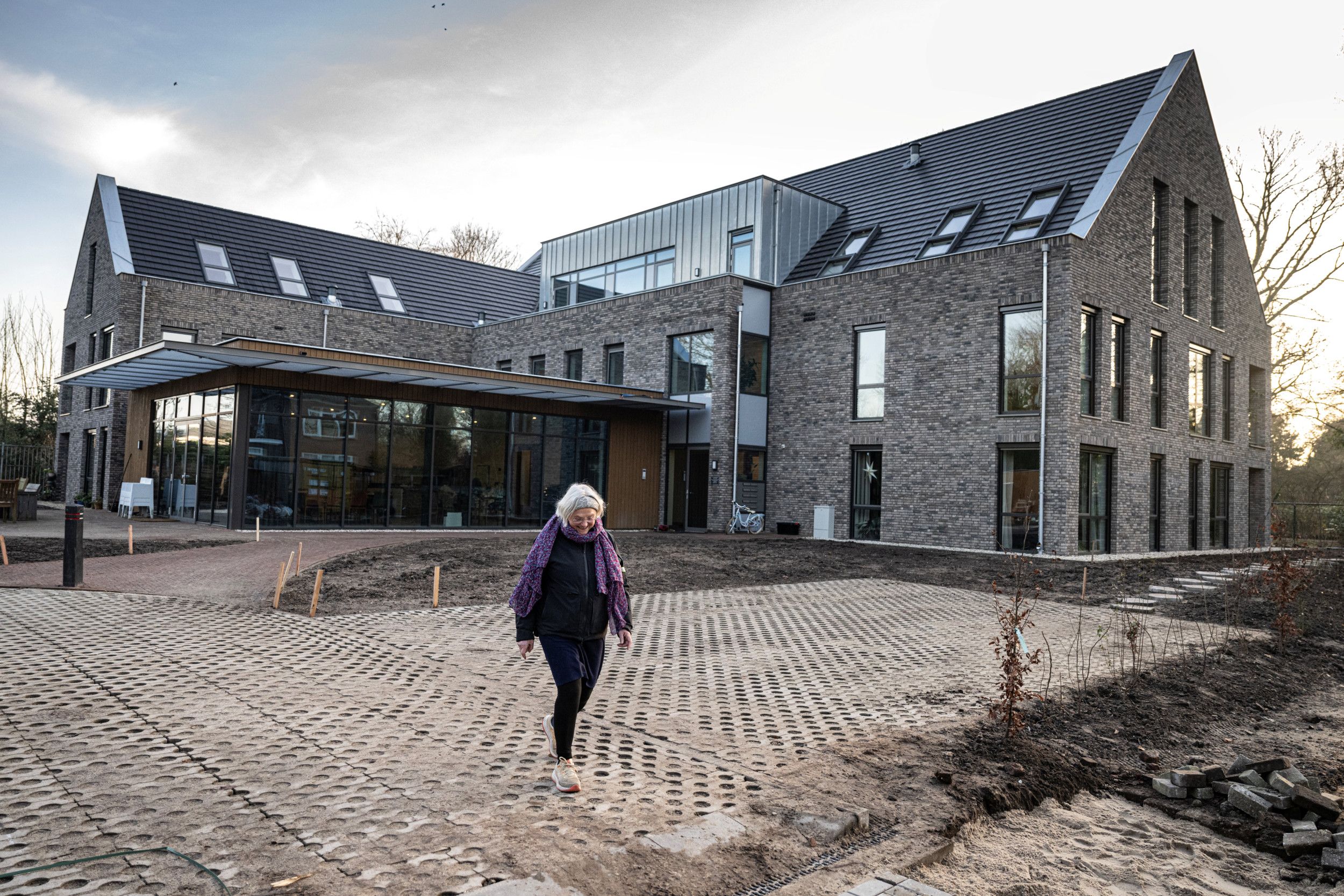 Oudere vrouw in donkere kleding met paarse sjaal loopt over de parkeerplaats van een appartementencomplex. 