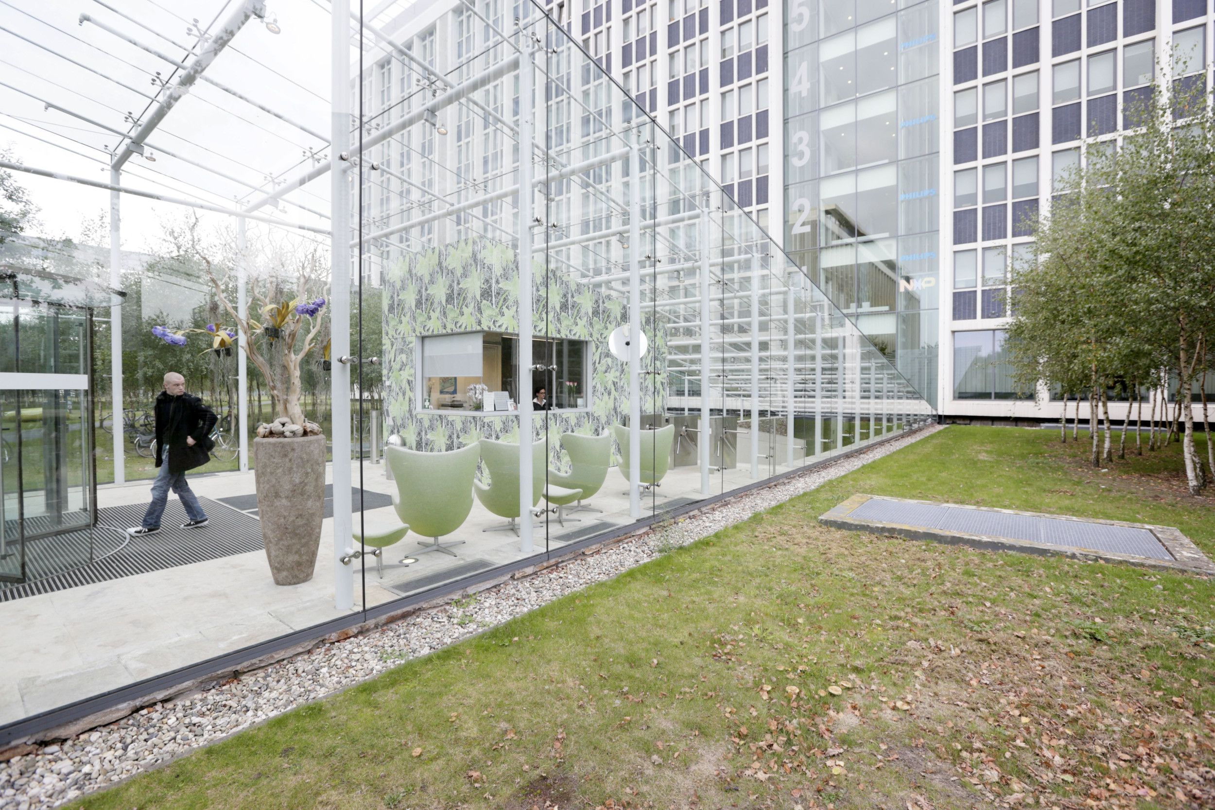 Een kaalgeschoren man met zwarte jas en spijkerbroek loopt door een glazen gang met daarin een balie waar een mevrouw zit. 