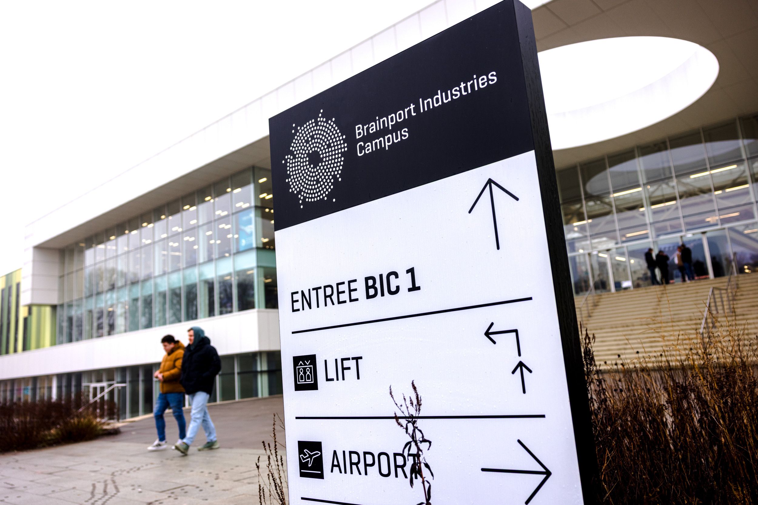 Twee jongeren lopen langs een bord waar onder andere de tekst Brainport Industries Campus op staat.  Het bord staat voor een groot gebouw met glazen gevel.