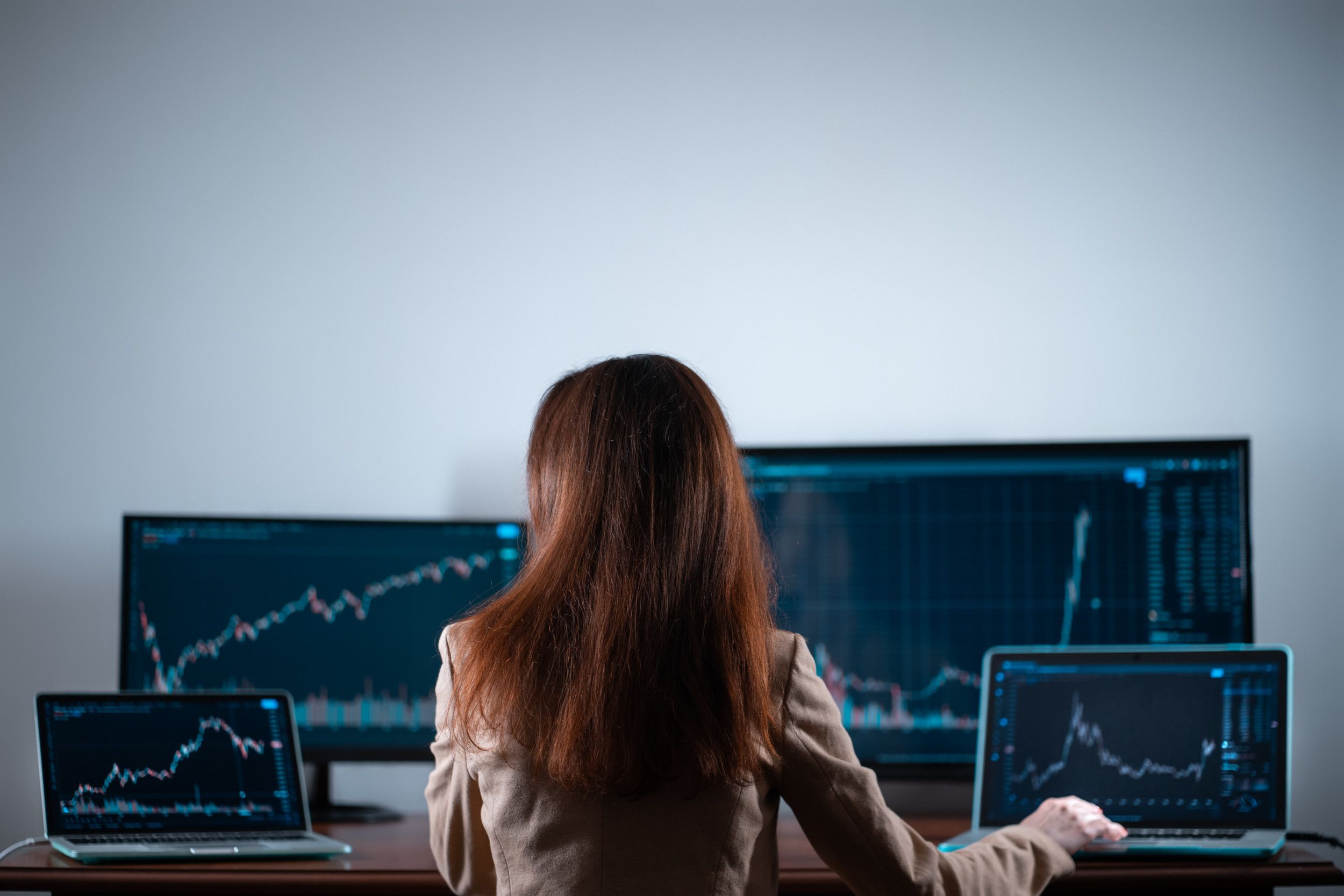 Een vrouw met lang bruin haar, op de rug gezien, werkt aan een bureau waar vier beeldschermen op staan. Op elk scherm prijkt een grafiek.