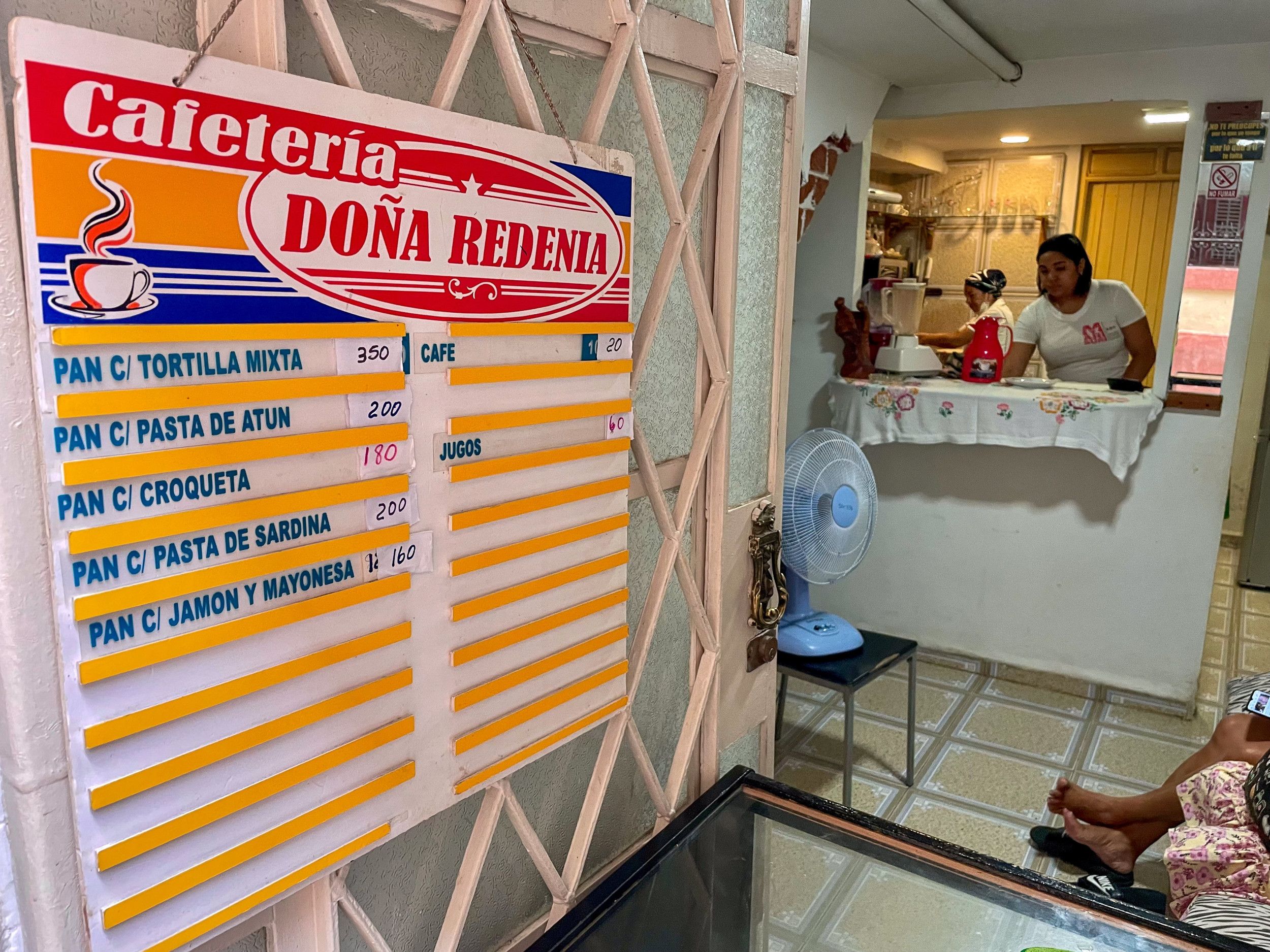 Een cafetaria aan huis in Havana. Achter de toonbank staan twee vrouwen. In de wachtruime staat een ventilator.