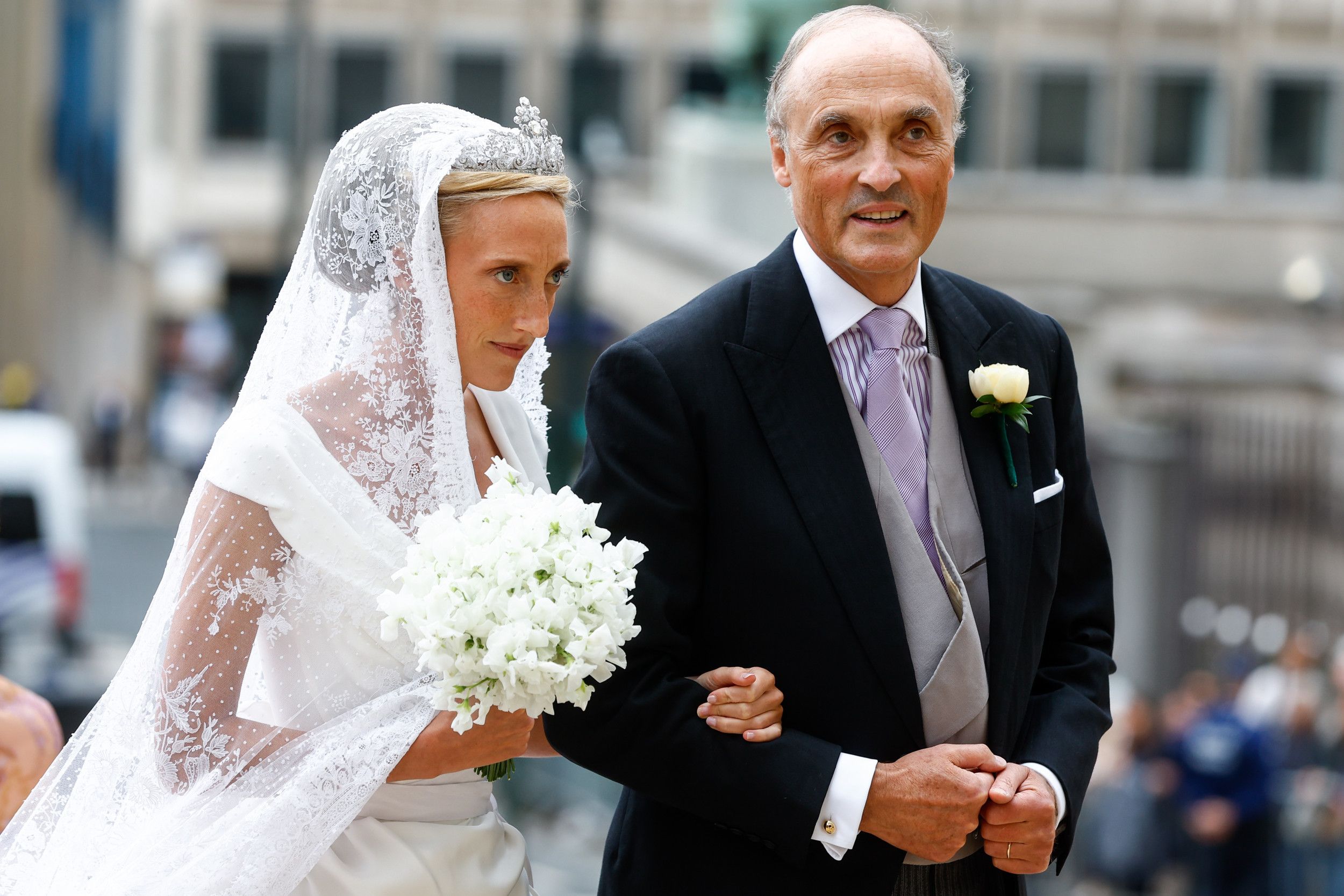 Bruid met lange sluier en wit boeket aan de arm van haar vader.