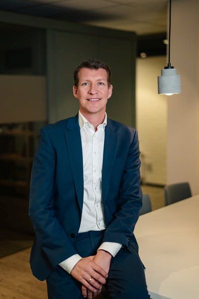 Een man in een blauw kostuum en wit overhemd zit met zijn handen over elkaar op een tafel in een kantoor.