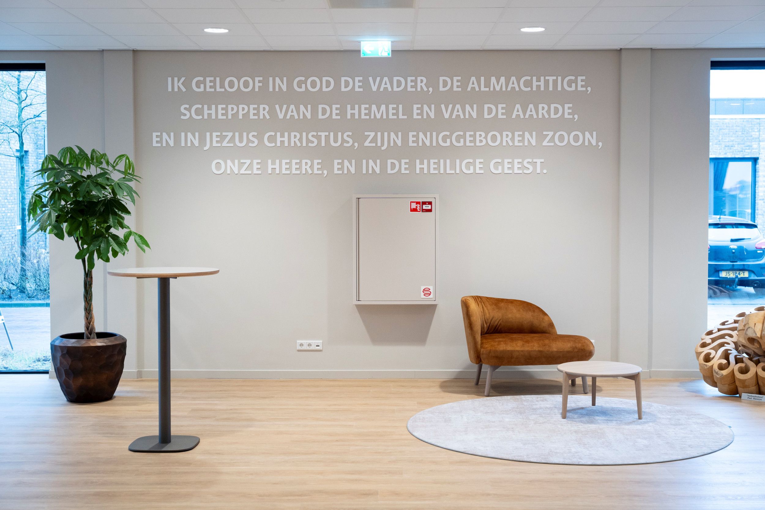 Tekst op de muur in de hal: „Ik geloof in God de Vader…”. Ervoor een modern vormgegeven stoel.